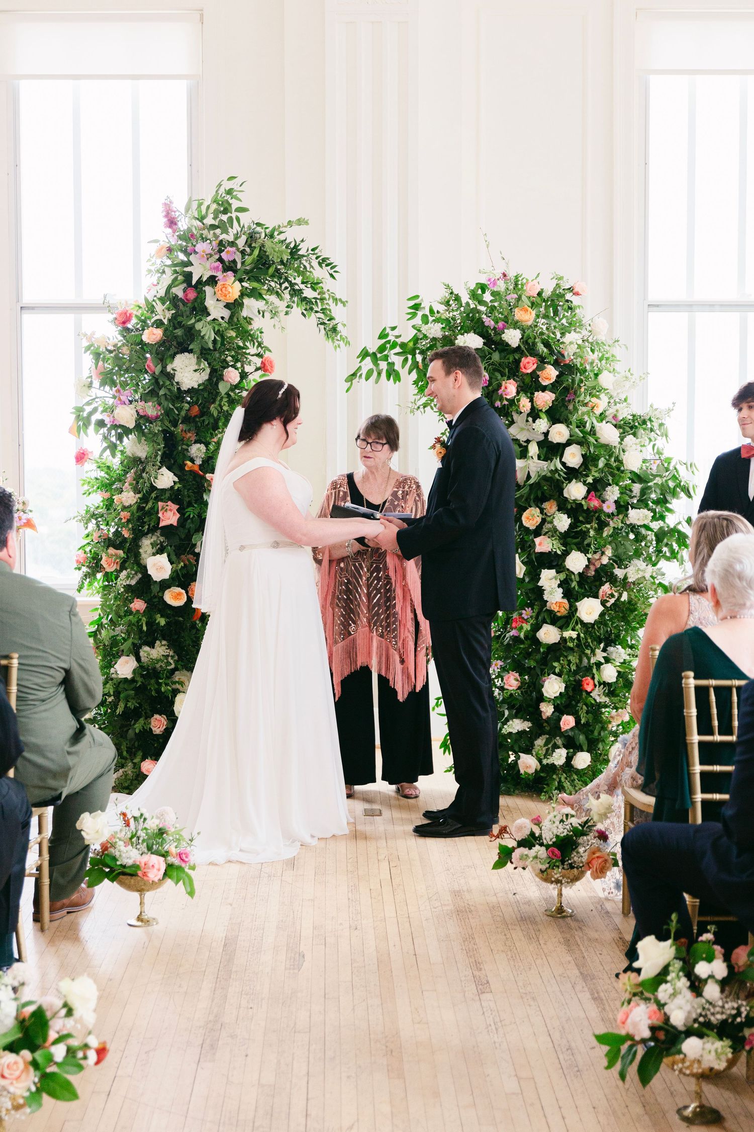 Bride and groom exchange vows beneath lush floral arches with pink and white blooms in a bright indoor wedding ceremony.