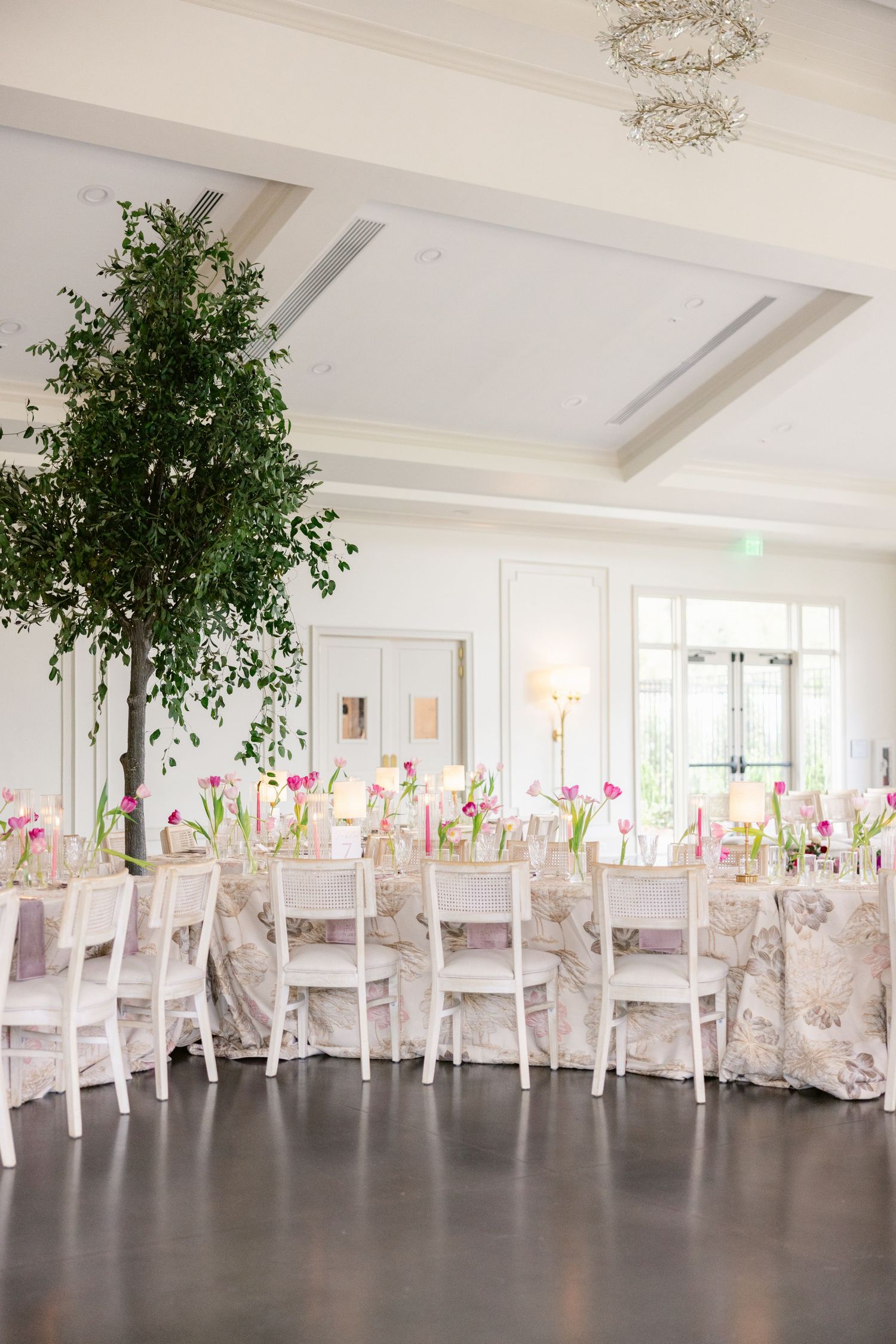 Spacious wedding venue with white chairs, floral tablecloths, and green potted trees.