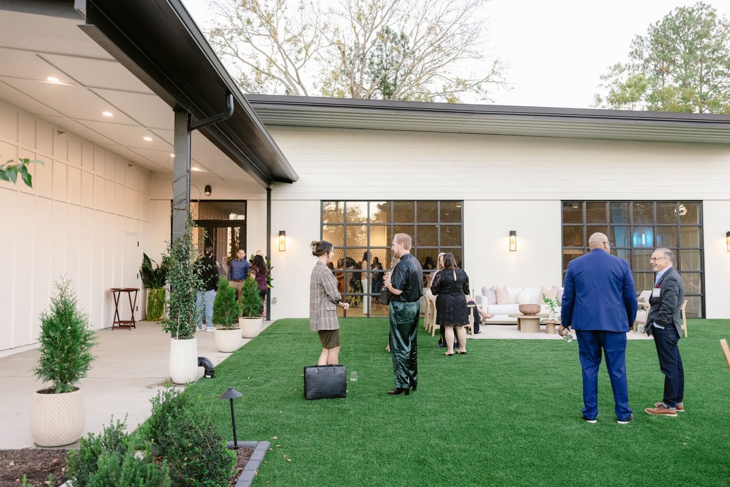 People stand outside a white building with glass windows on a grassy lawn during an outdoor gathering.