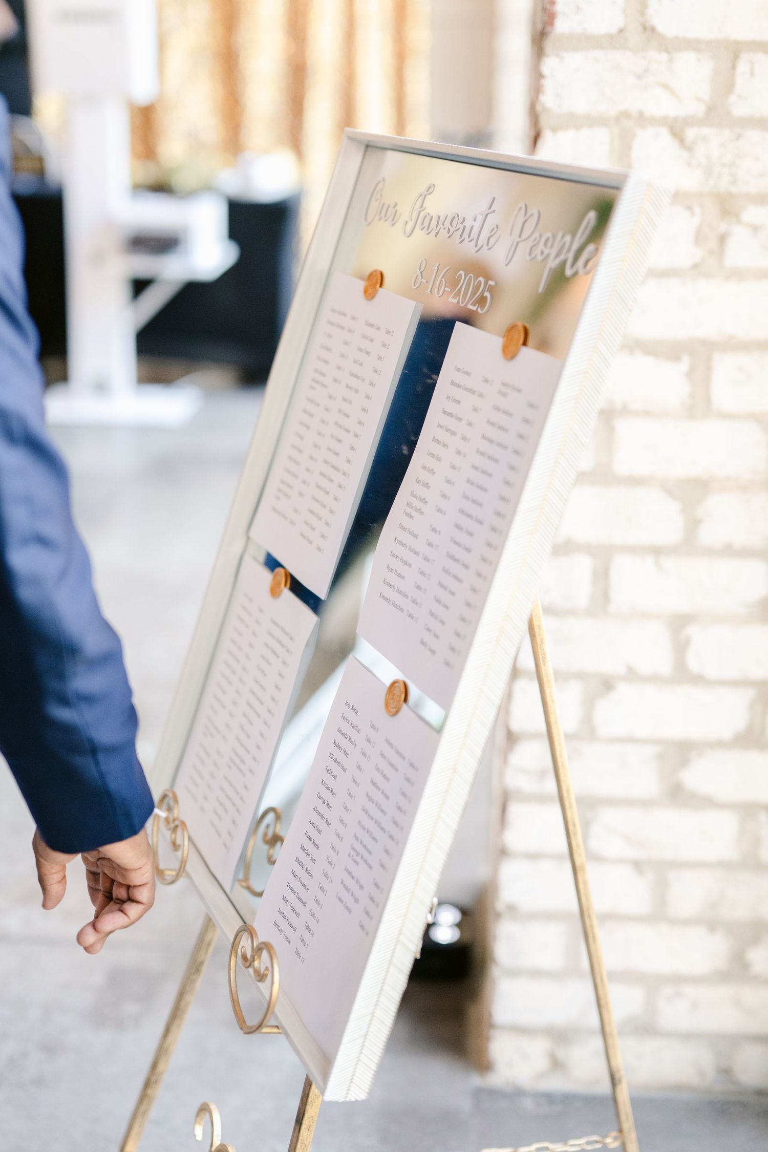 Wedding seating chart displayed on white easel with elegant calligraphy and gold accents against white brick wall.