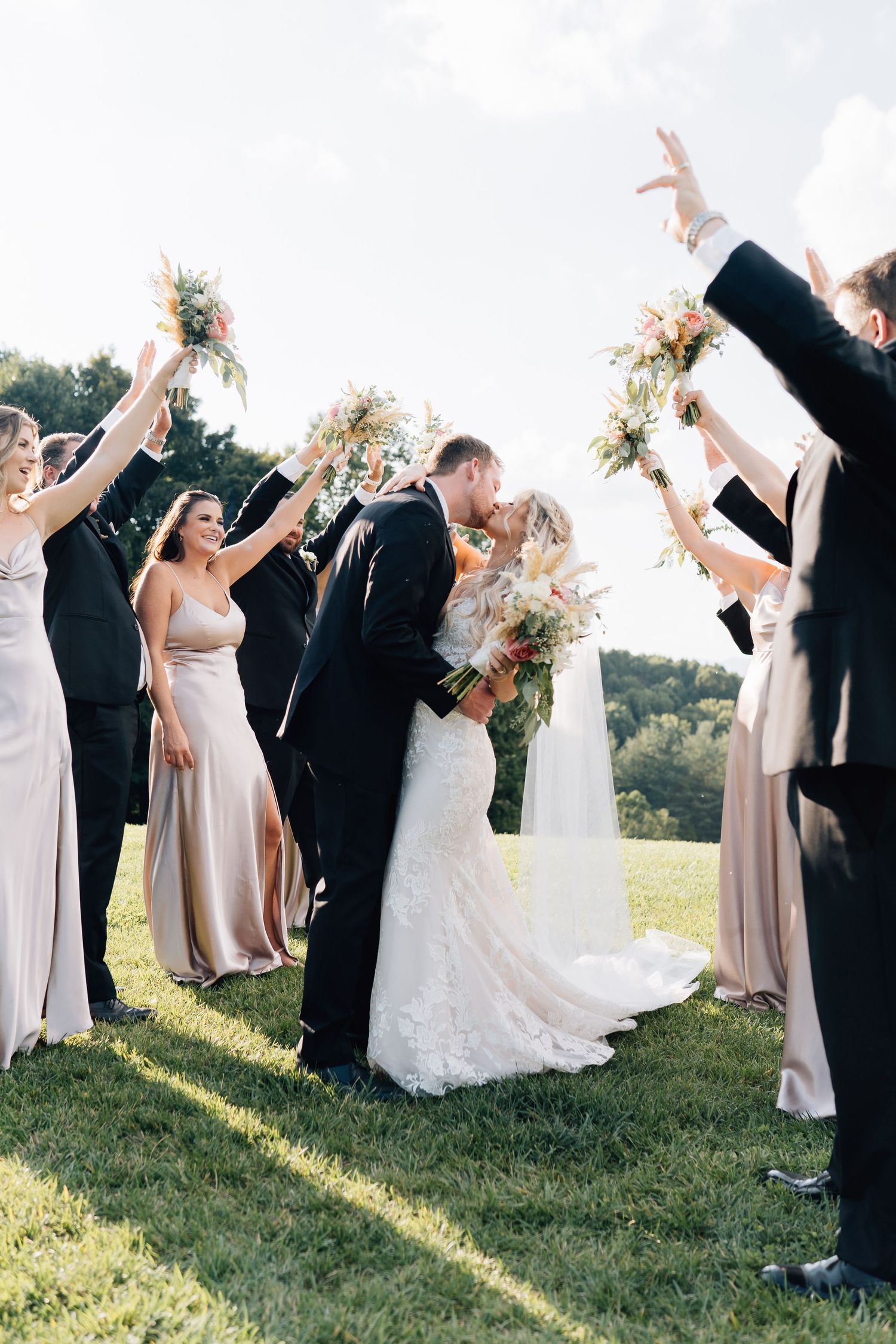Wedding party celebrates on a sunny lawn as bridesmaids in blush gowns hold bouquets high while couple shares a kiss.