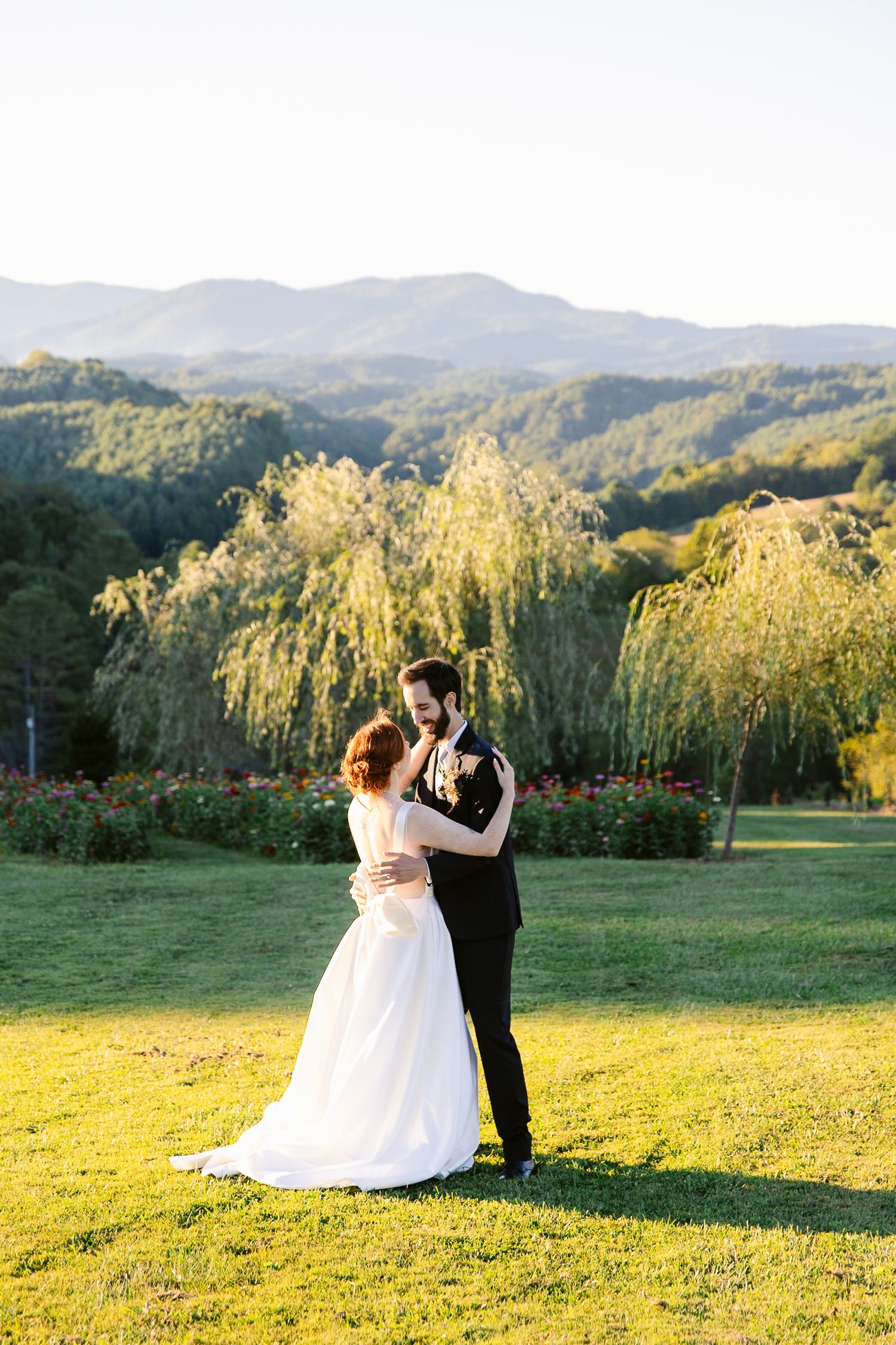 A couple shares their first dance at sunset with mountains and weeping willows in the background.
