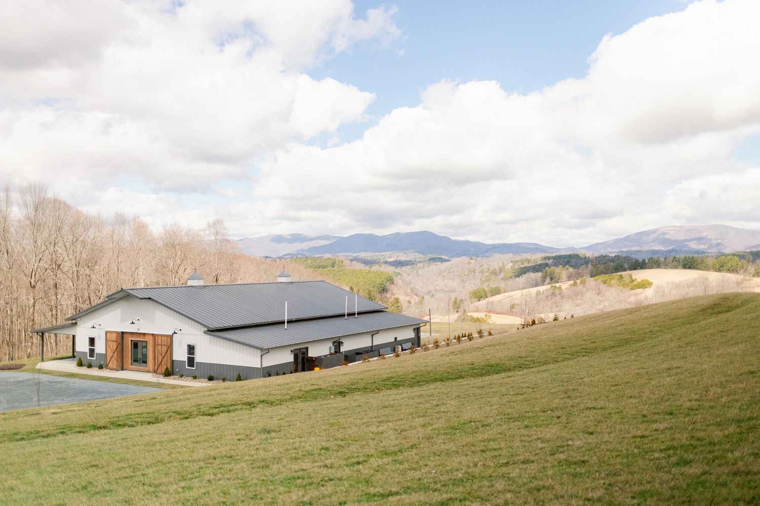 A panoramic view of rolling green hills with farm buildings and mountains in the distance under a cloudy sky.