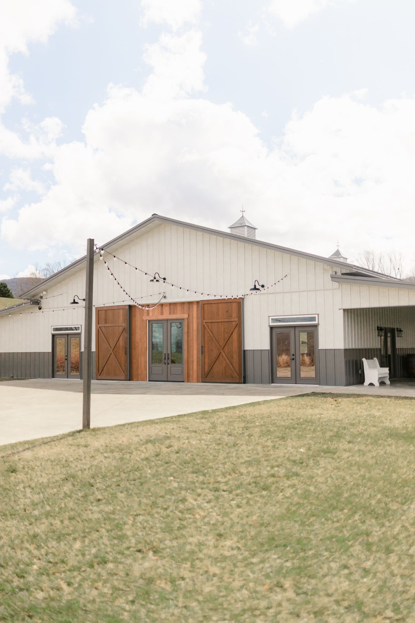 Long white barn-style building with wooden sliding doors and windows set against a grassy hillside on a cloudy day.