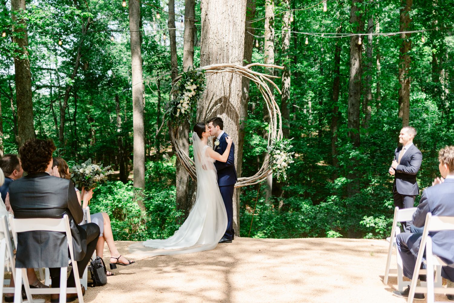 A panoramic view of a forest wedding ceremony with guests seated on wooden benches among tall trees.