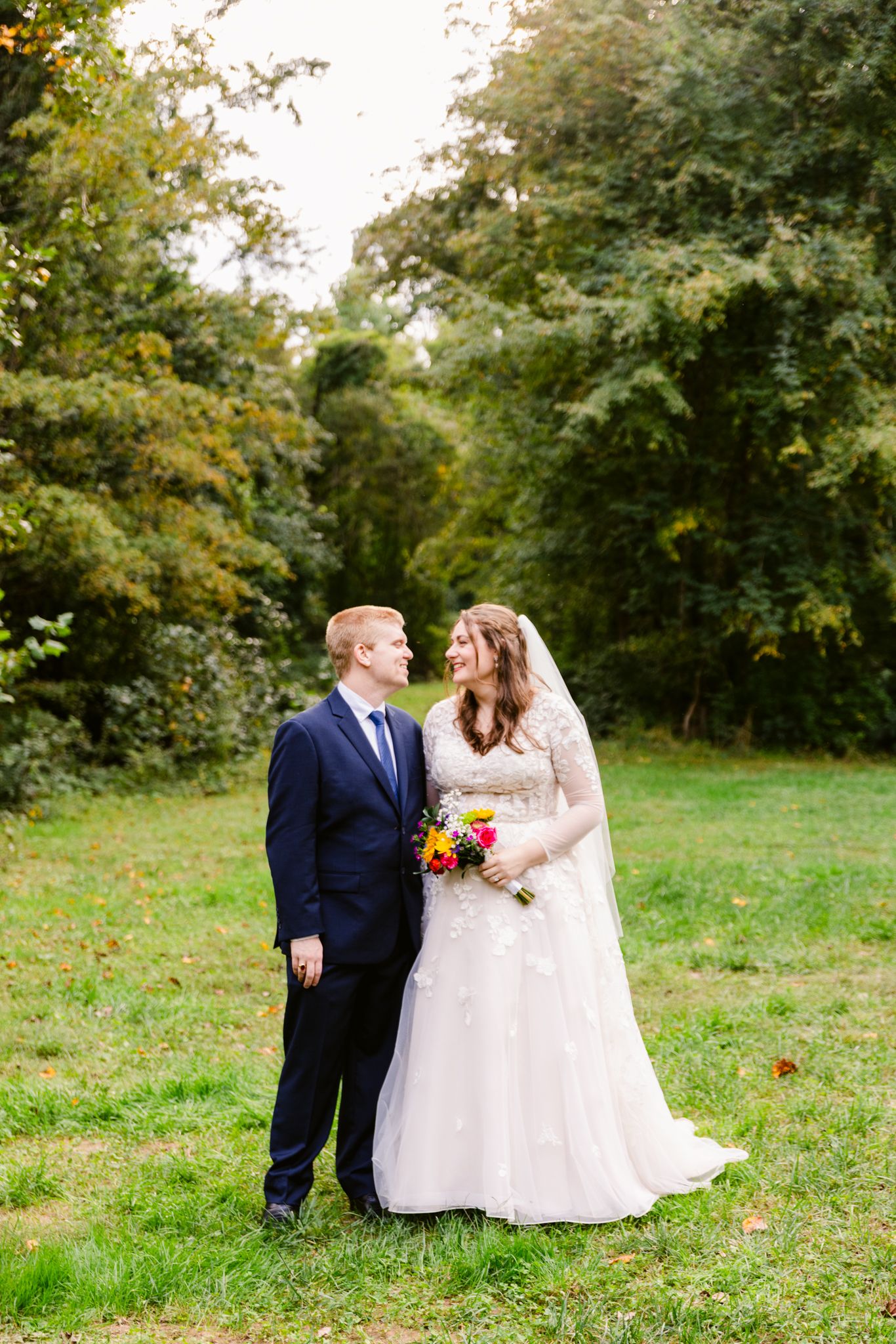 A romantic outdoor wedding scene with a couple in formal attire standing together on green grass surrounded by trees.