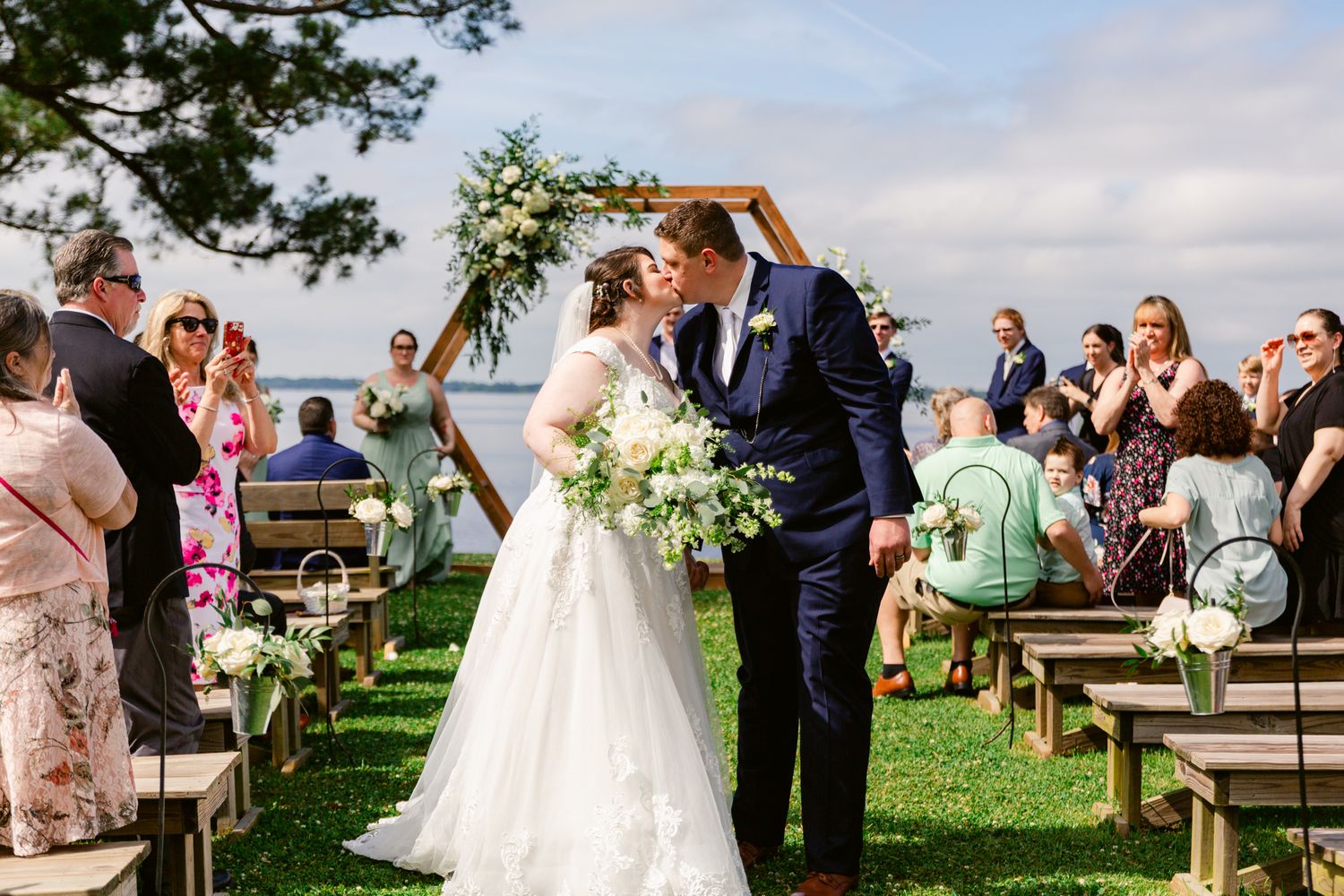 A wedding ceremony takes place on a grassy hilltop with wooden benches and guests watching the newlyweds share a moment.