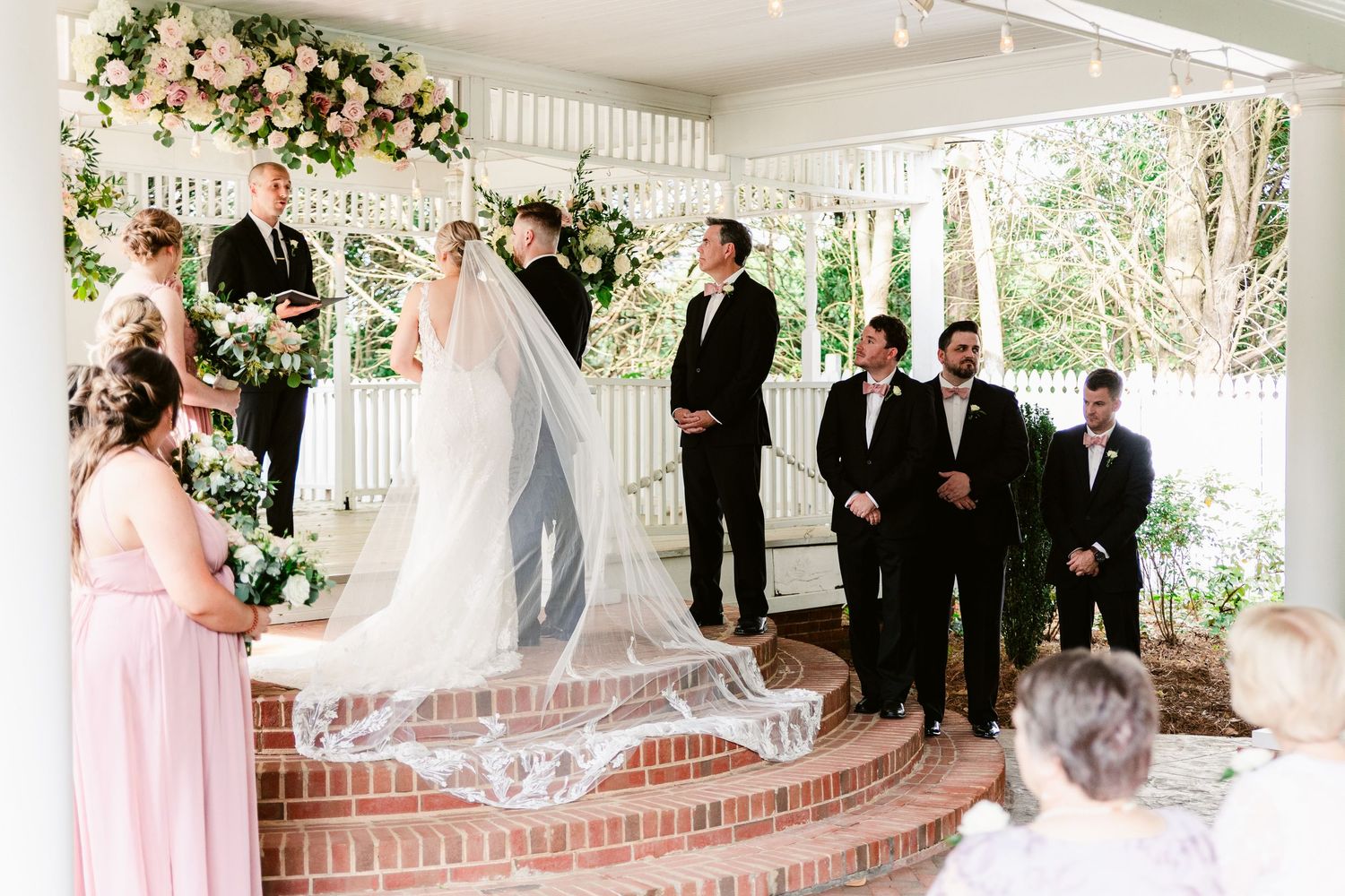 A panoramic view of an outdoor wedding ceremony under a white gazebo decorated with floral arrangements and greenery.