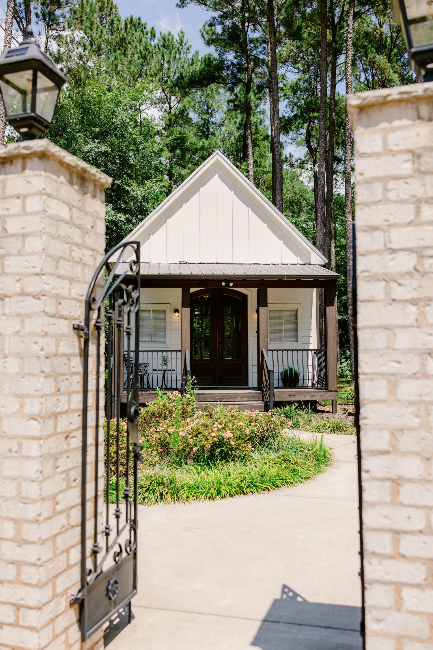 White cottage-style house with stone pillars and wrought iron gate entrance surrounded by lush greenery and pine trees.