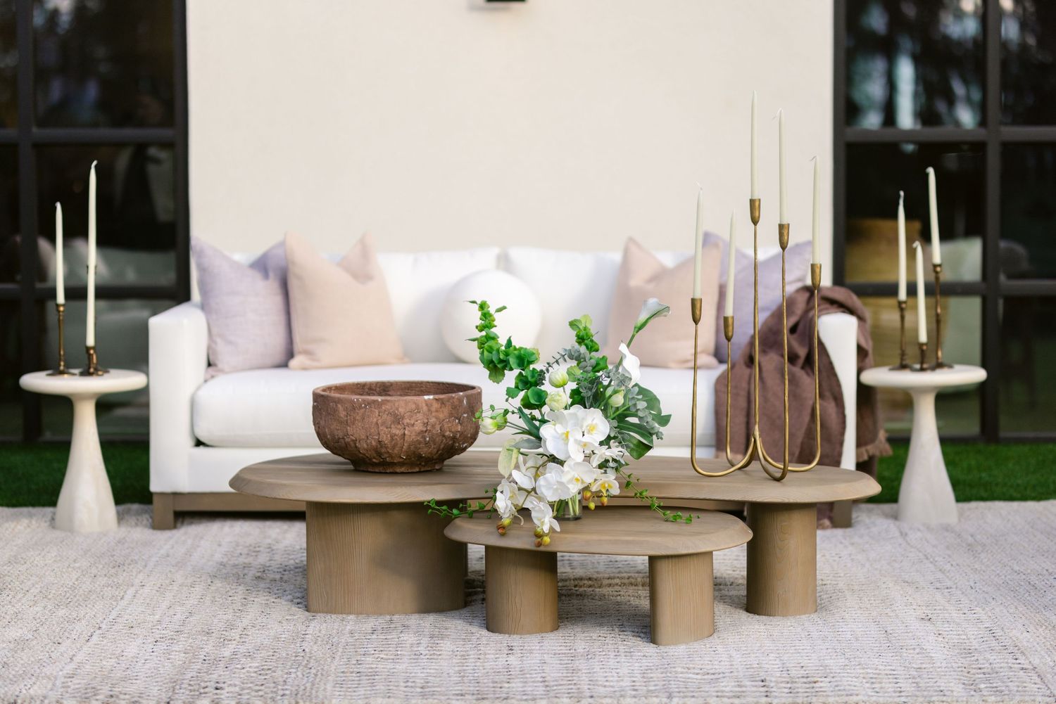 Modern outdoor lounge area with neutral-toned cushions, decorative pillows, and potted plants on a paved patio.