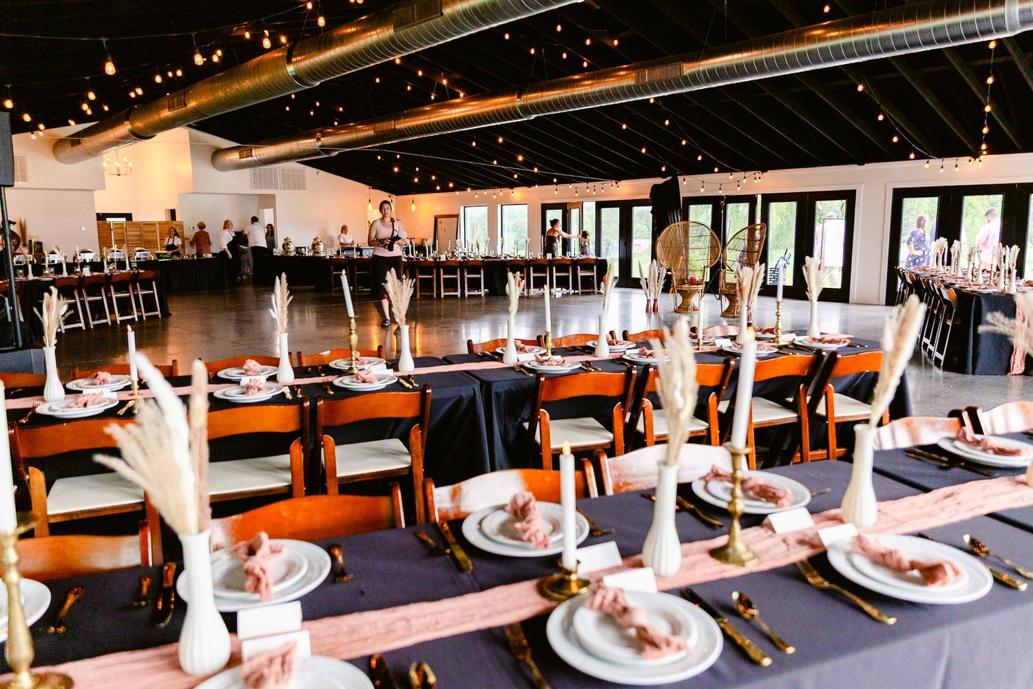 Long banquet tables with white linens and place settings are arranged in an elegant dining room with large windows.