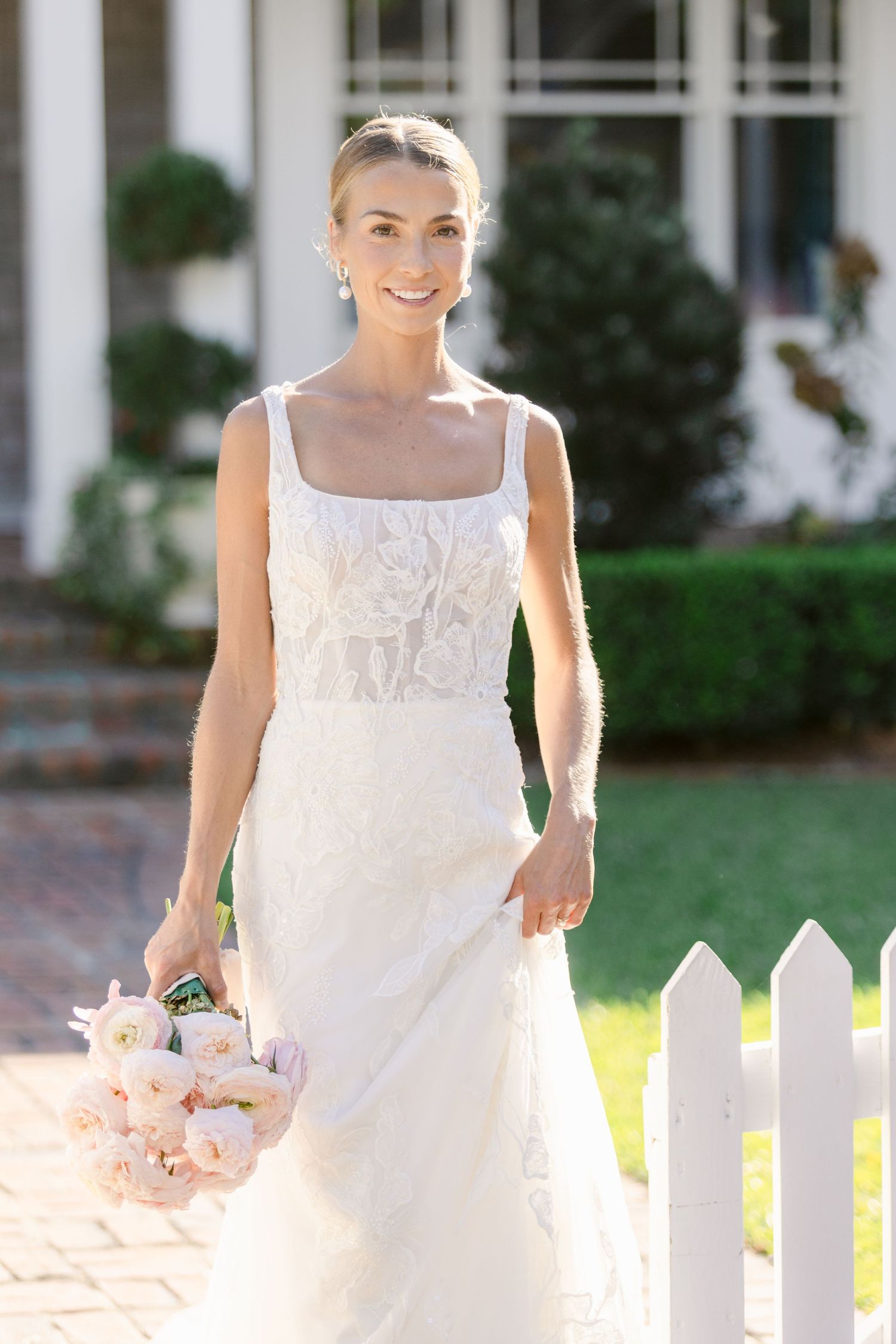 Elegant bride in lace wedding gown holding blush bouquet, smiling outdoors near white picket fence.