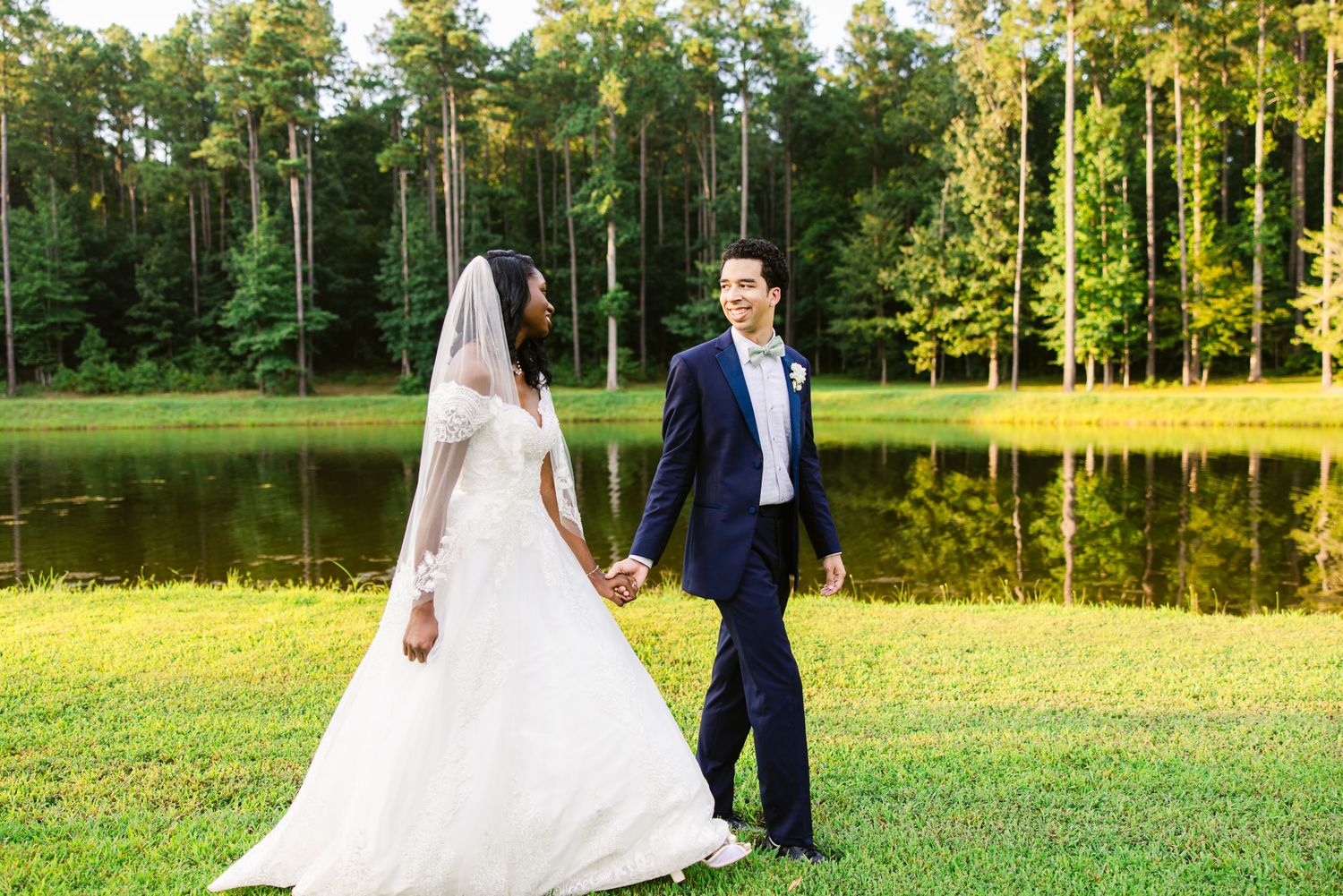 Wedding couple walking through a sunlit meadow surrounded by tall trees.