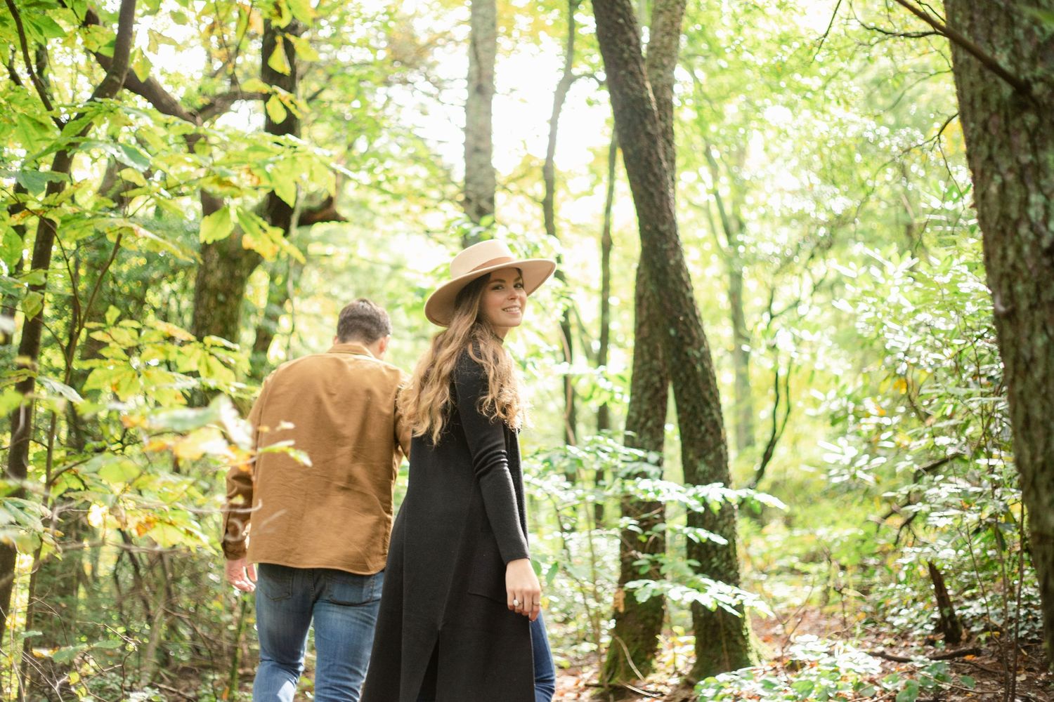Couple walking through a sunlit forest path in casual fall clothing.