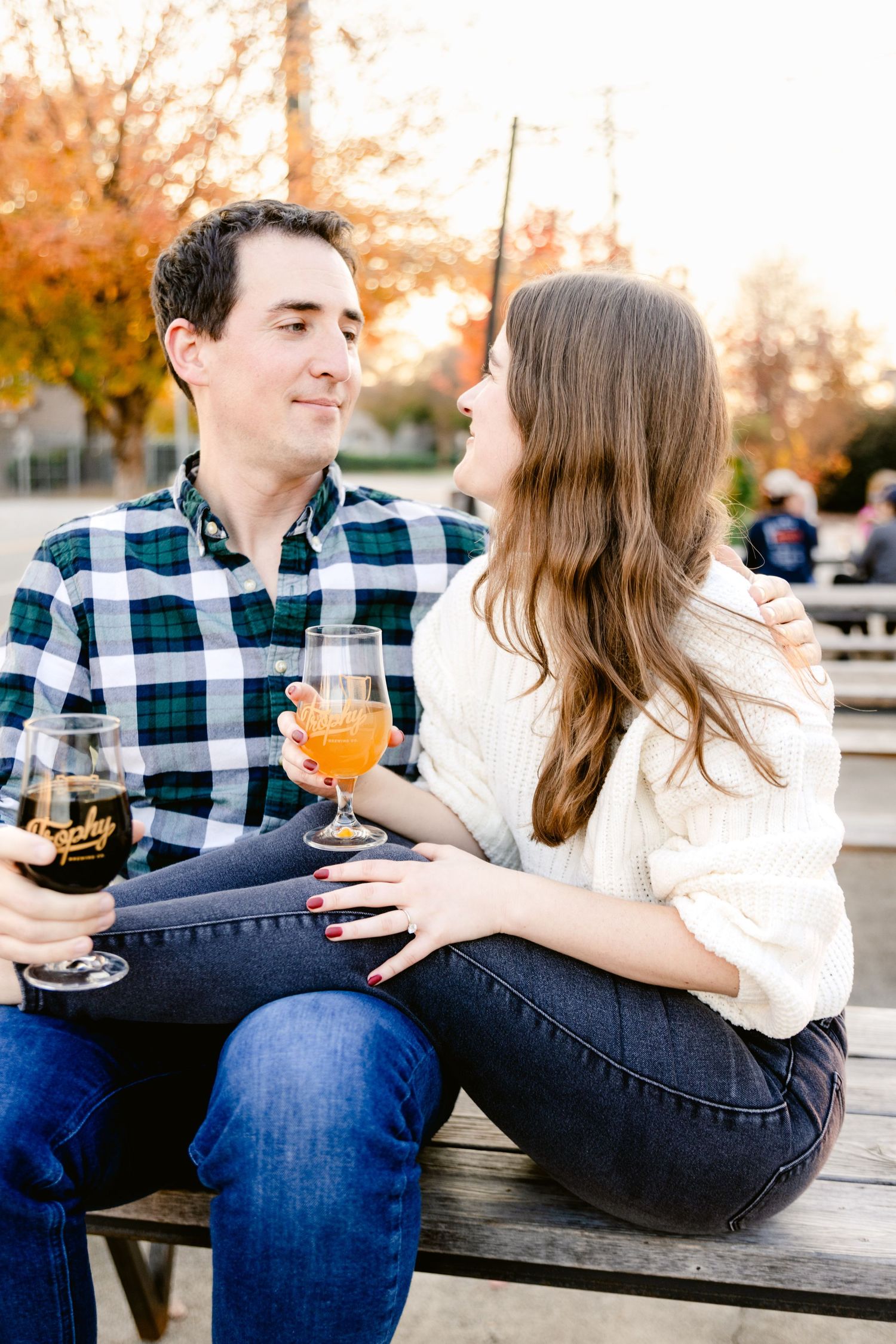 A couple enjoys cocktails together on a wooden bench during autumn sunset with golden foliage in background.