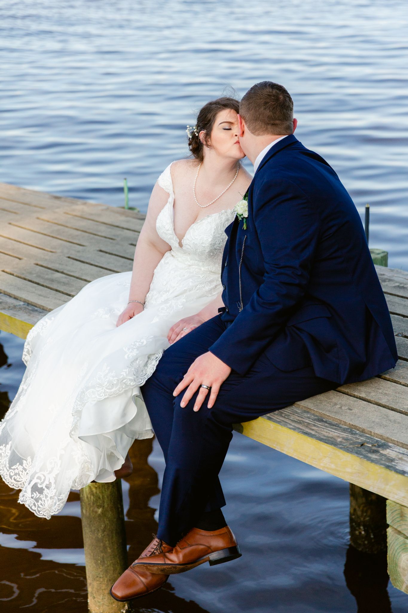 A romantic wedding couple sits together on a wooden dock by the water in formal attire.