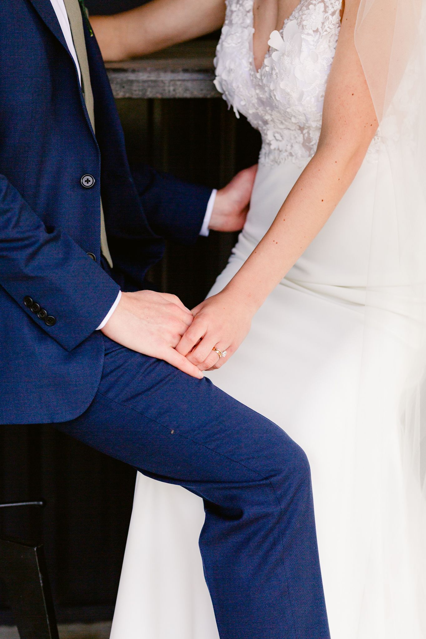 Close-up details of bride in white lace dress and groom in navy suit sitting together.