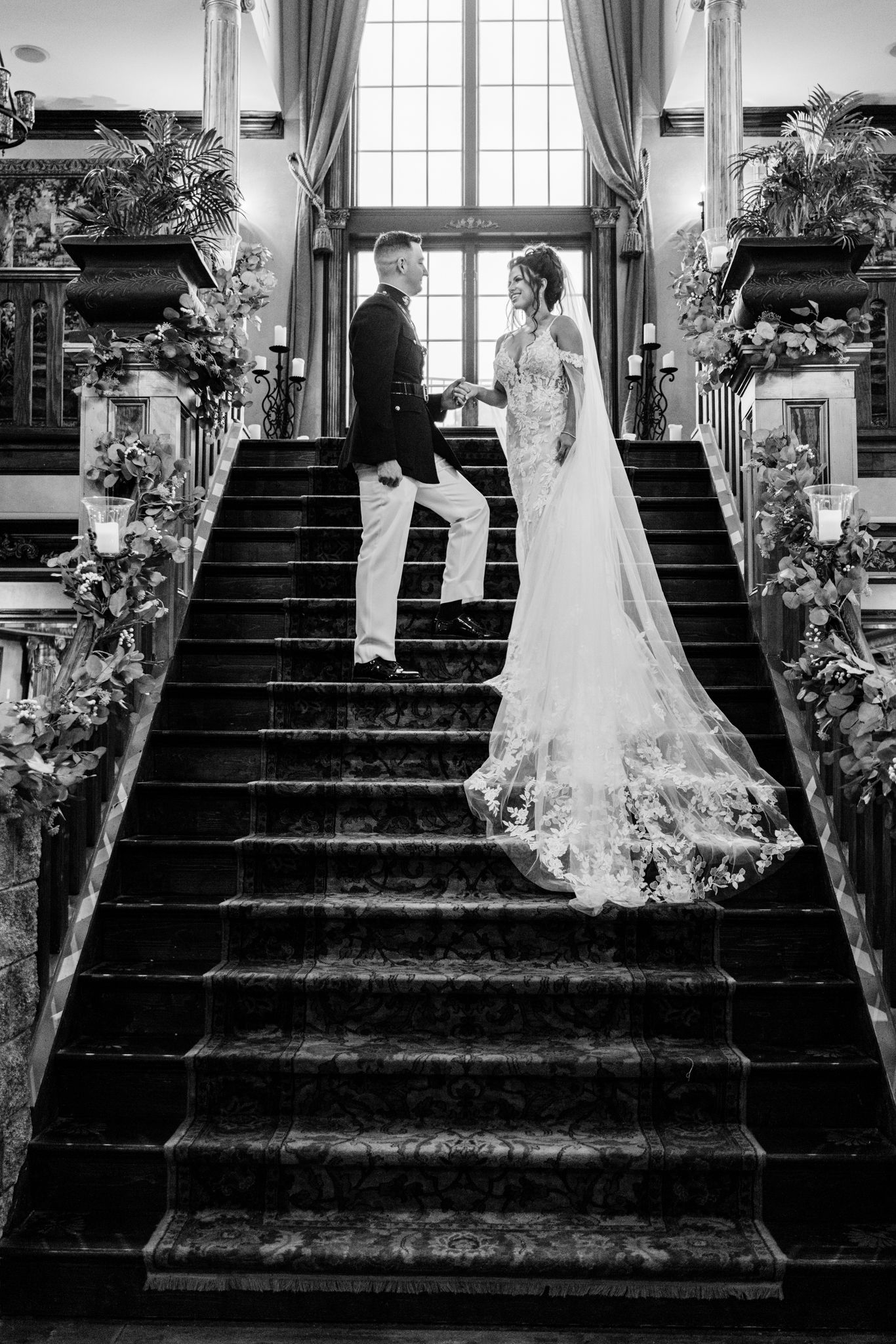A romantic black and white wedding photo of a couple standing on an elegant staircase adorned with floral arrangements.