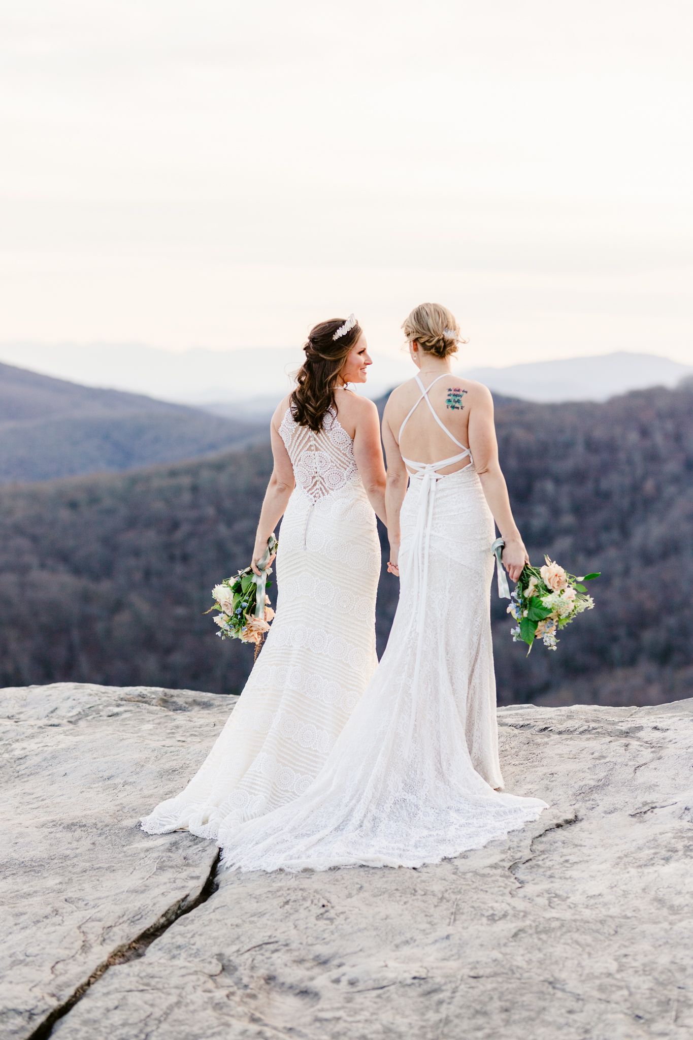 Two people in white wedding dresses sit together on a rocky mountain overlook with scenic mountain views.