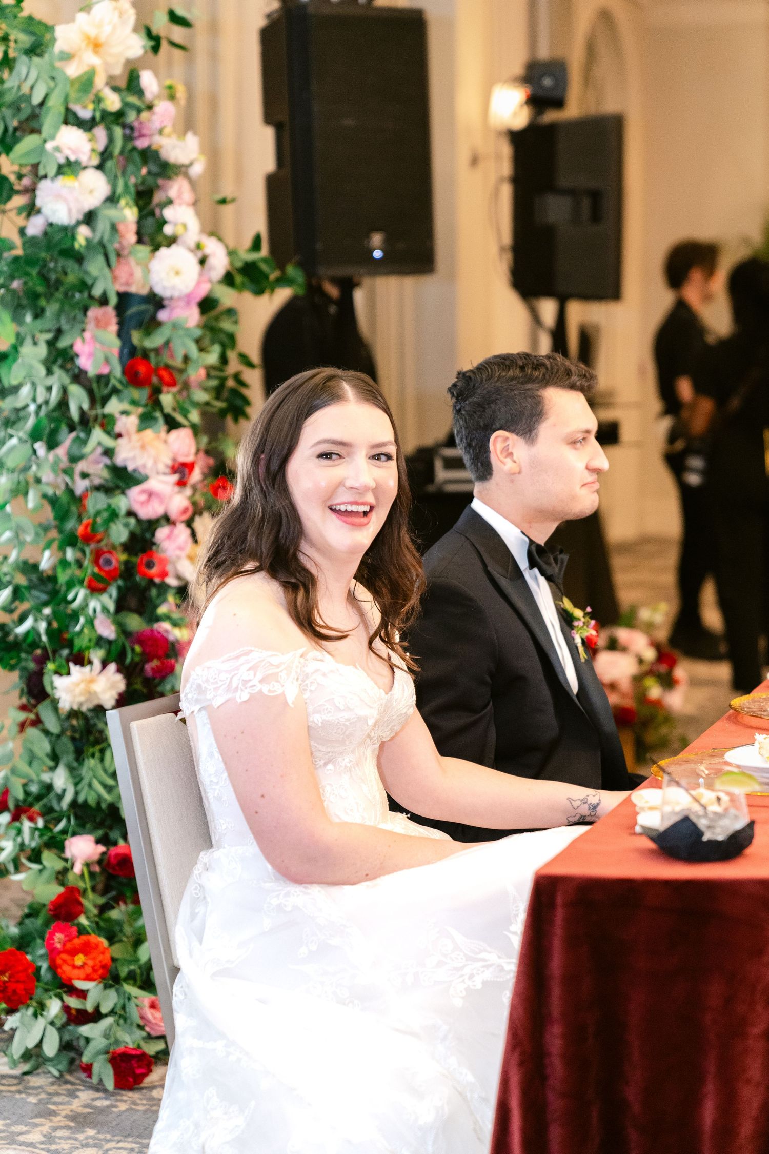 An elegant wedding reception setup with floral decorations and burgundy tablecloths in a bright indoor venue.