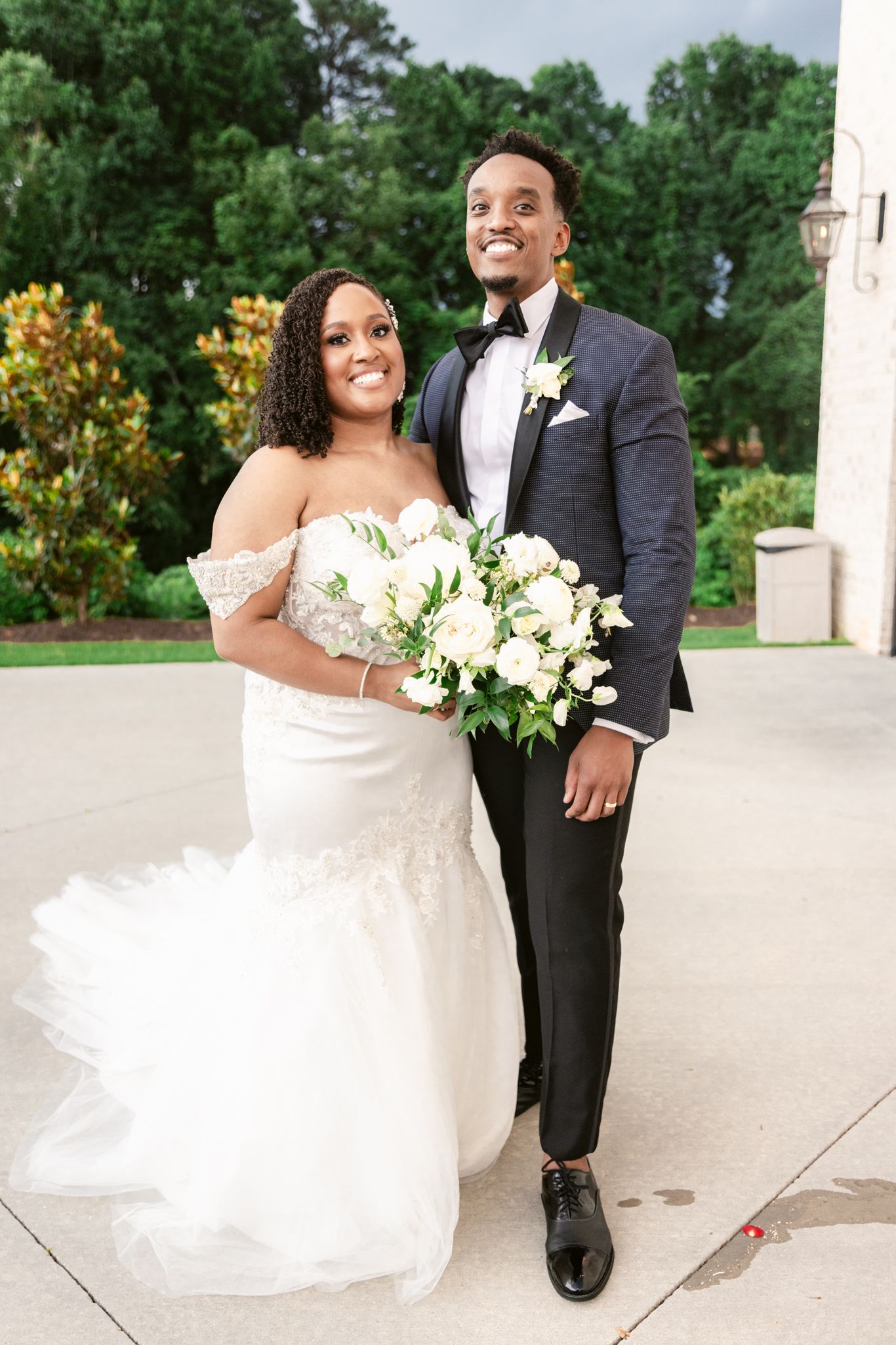 A bride in an off-shoulder white gown holds a white floral bouquet while standing next to a groom in a black tuxedo outdoors.