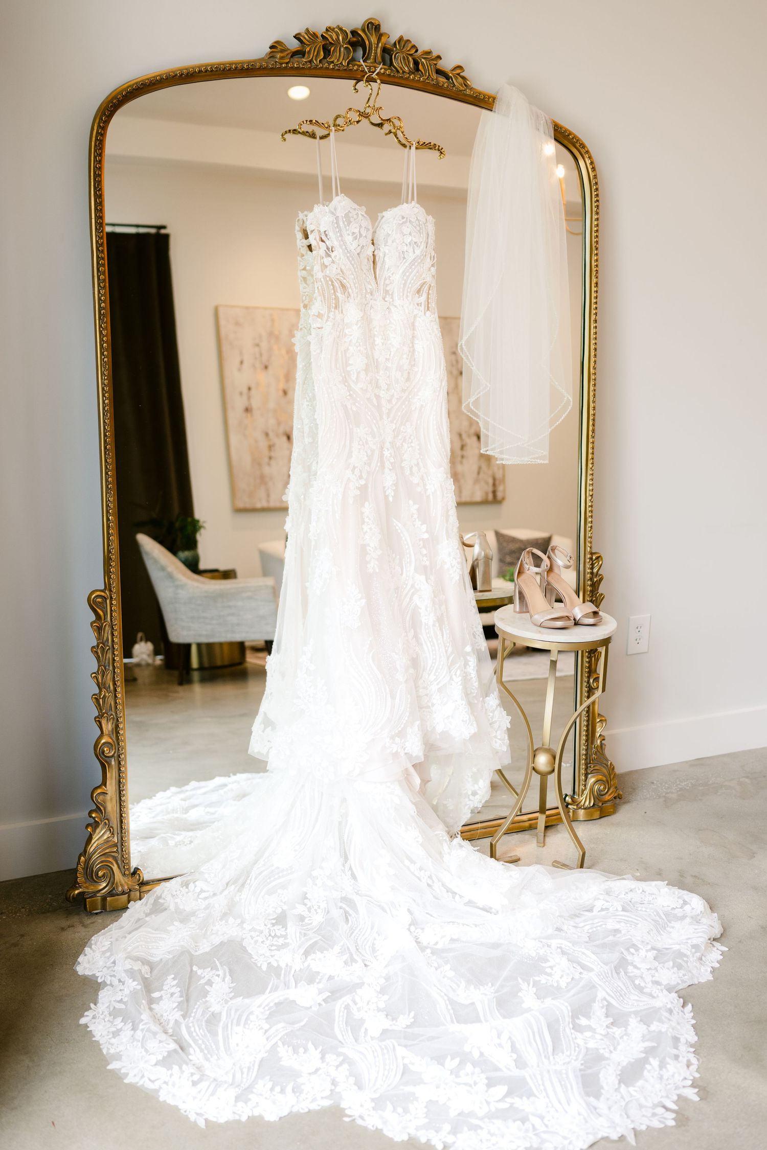 Stunning lace wedding dress with long train displayed on hanger in front of ornate gold mirror.