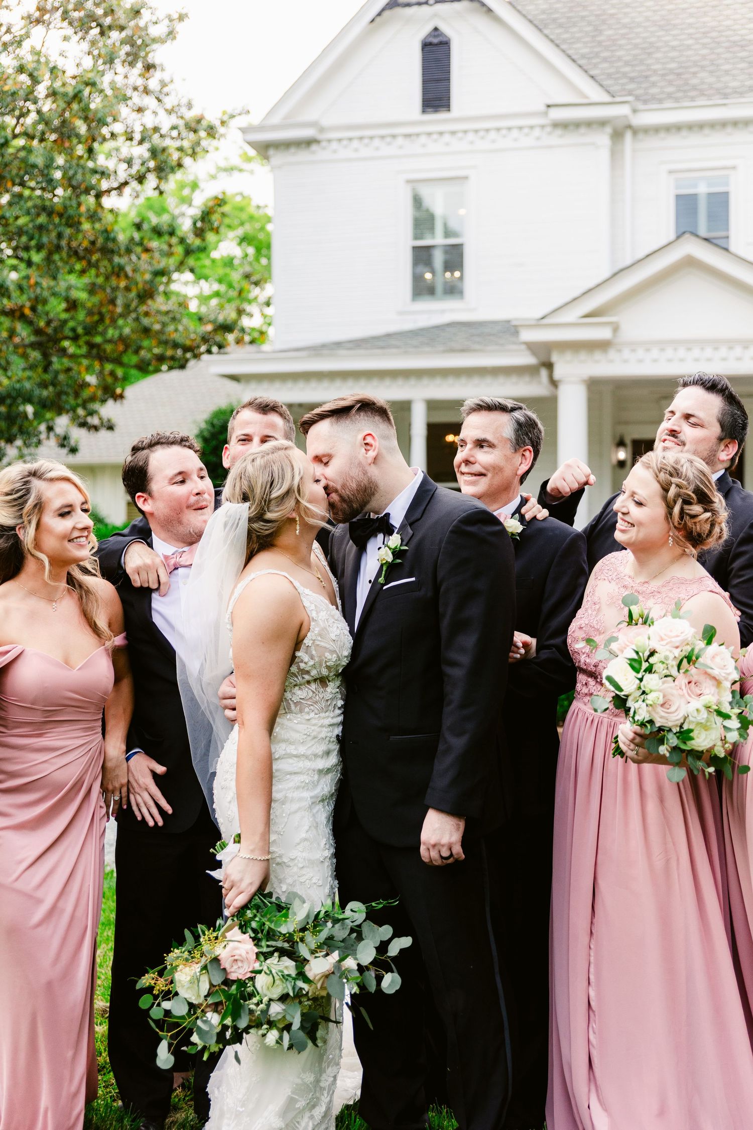 Wedding party in black suits and pink bridesmaid dresses poses together in front of white house.