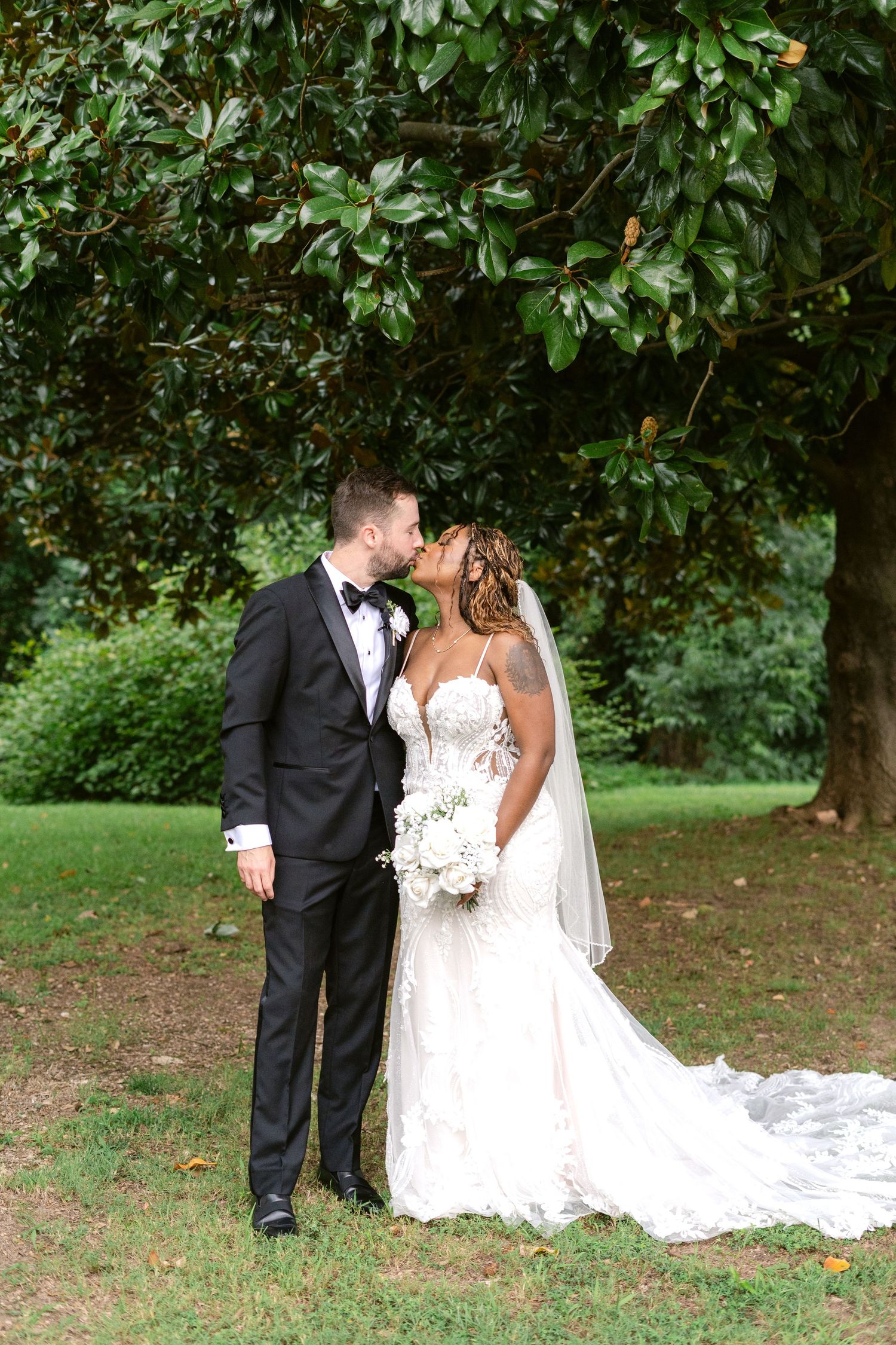 A romantic wedding moment captured under lush green trees as the couple shares an intimate kiss.