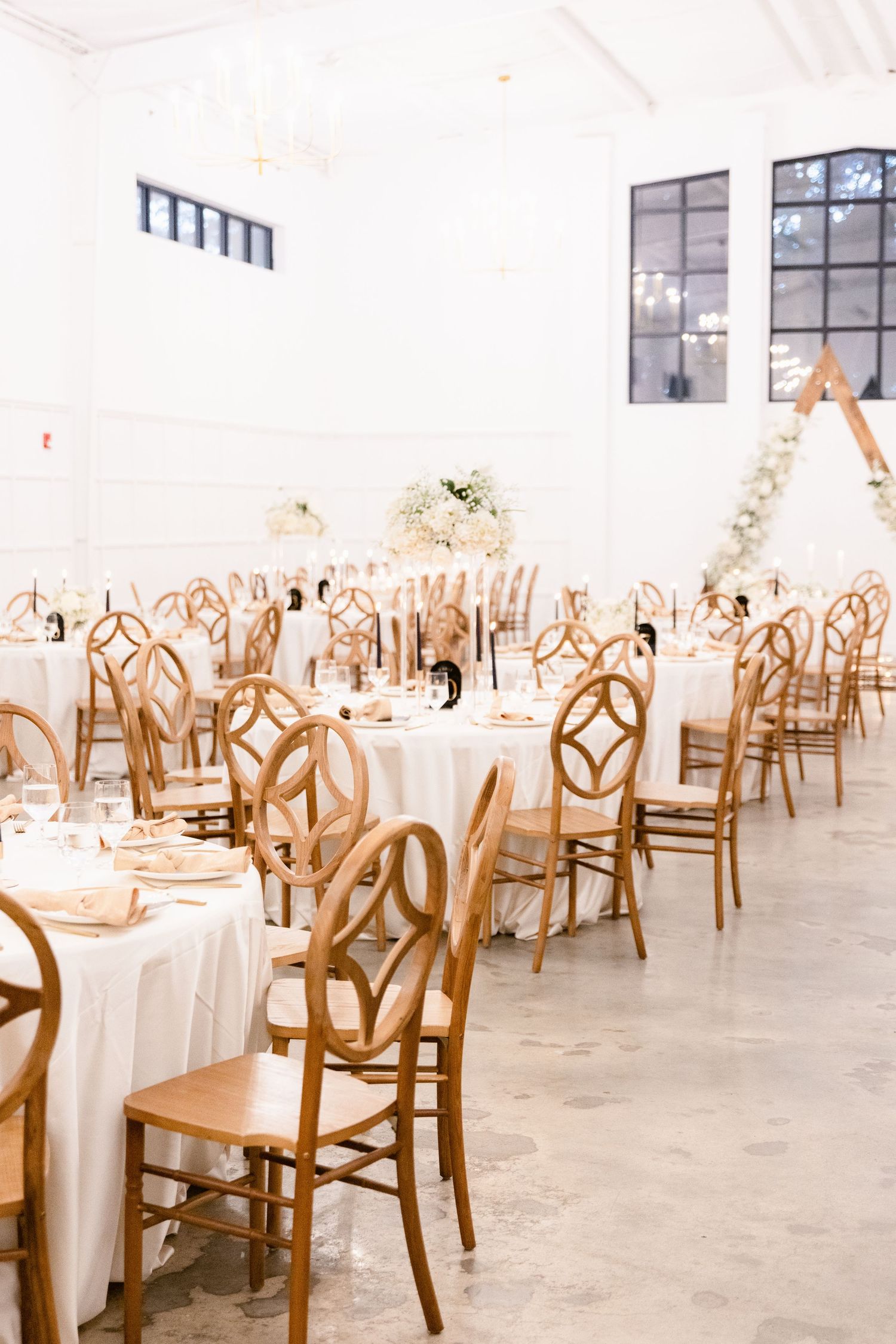 Large white reception hall with wooden chairs and round tables set for an event.