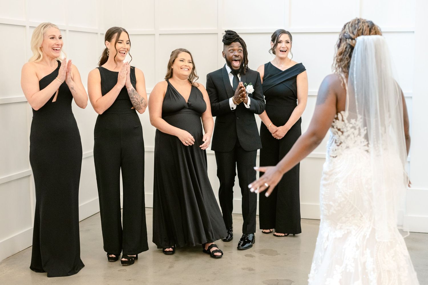 Wedding party wearing elegant black attire reacts with joy as they see bride in white gown and veil.