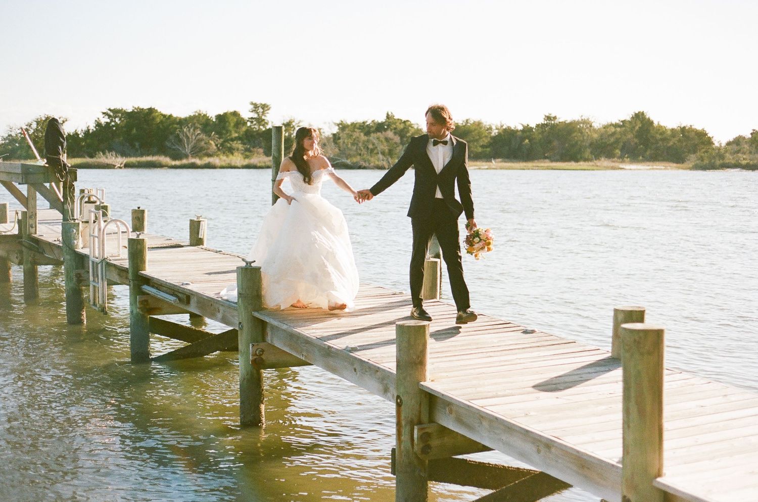 A wedding couple walks along a wooden dock by the water with their wedding party following behind them.