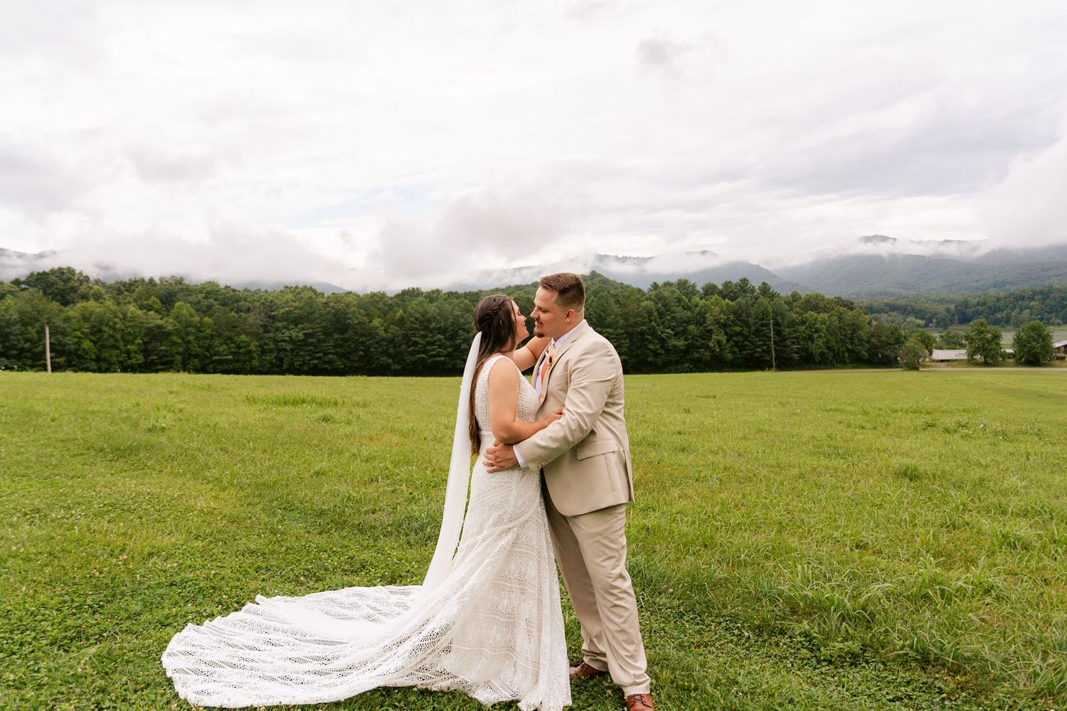 A couple embraces in a lush green field with mountains and forest in the background on their wedding day.
