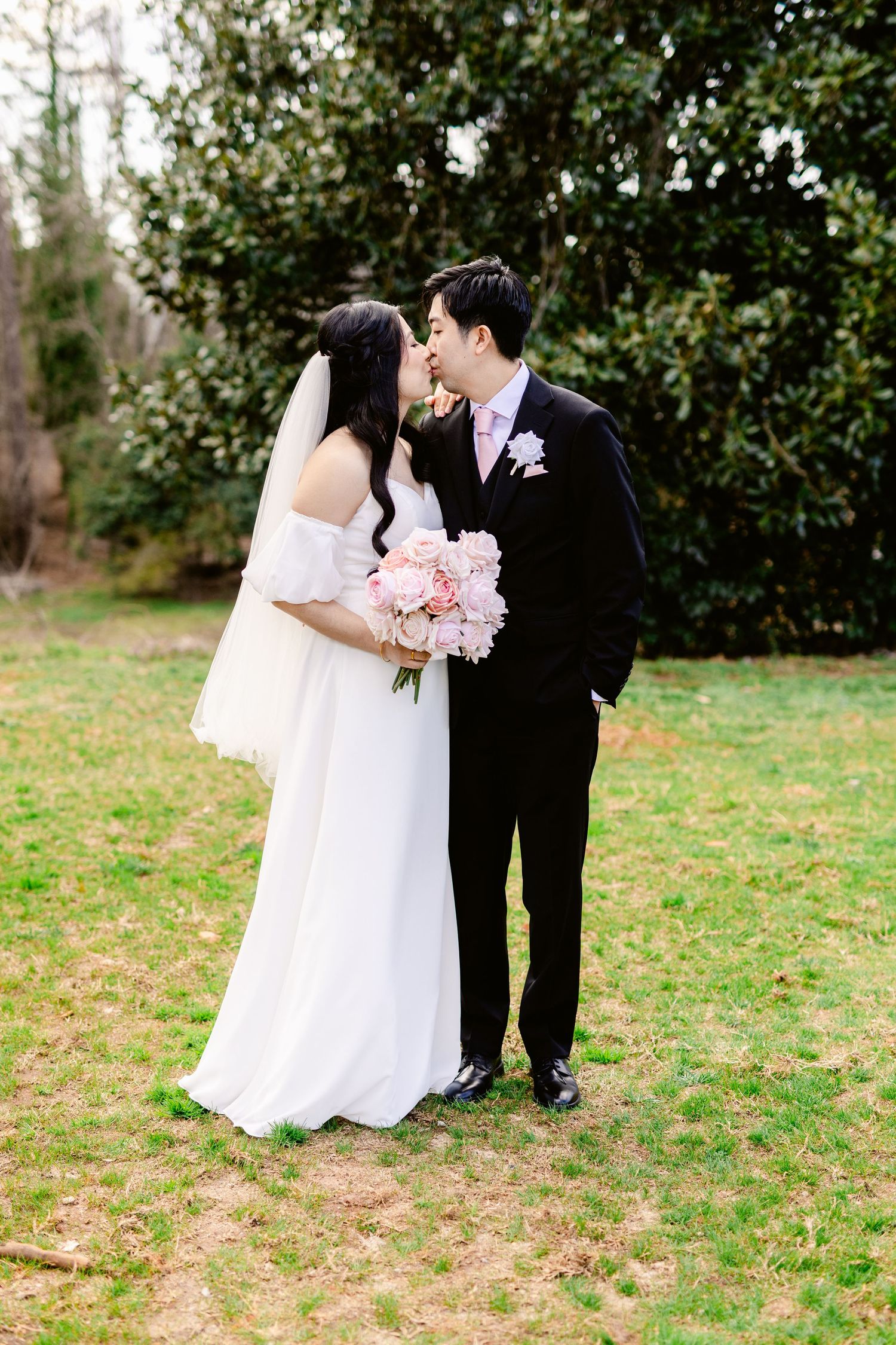 A romantic wedding couple shares a tender moment in a garden setting with a pink bouquet and elegant attire.