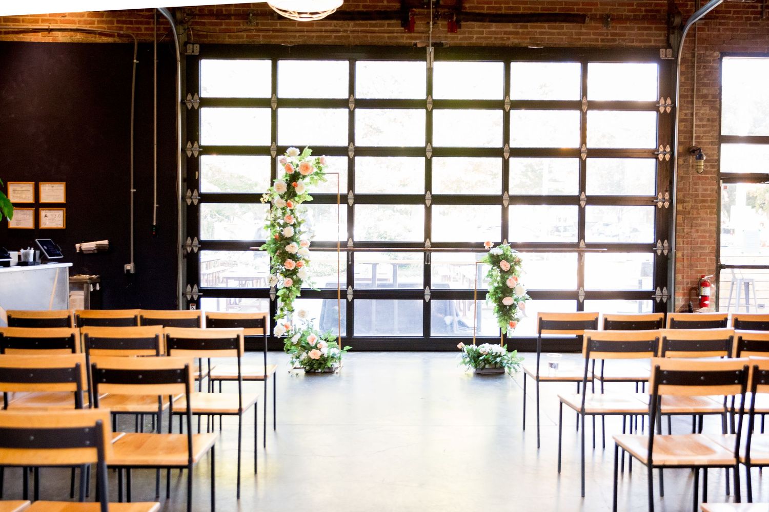 Modern industrial wedding ceremony space with wooden chairs and floral arrangements along the aisle near a glass garage door.