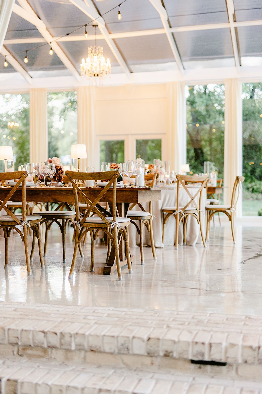Light-filled reception hall with wooden cross-back chairs and crystal chandeliers creating an airy, romantic atmosphere.