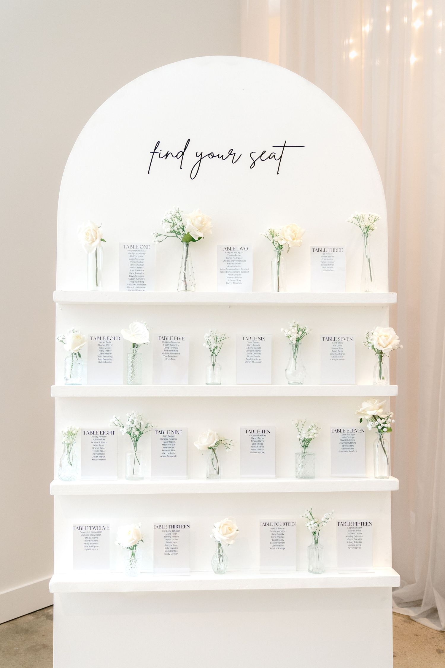 White seating chart display with 'find your seat' text and small bud vases holding white flowers on tiered shelves.