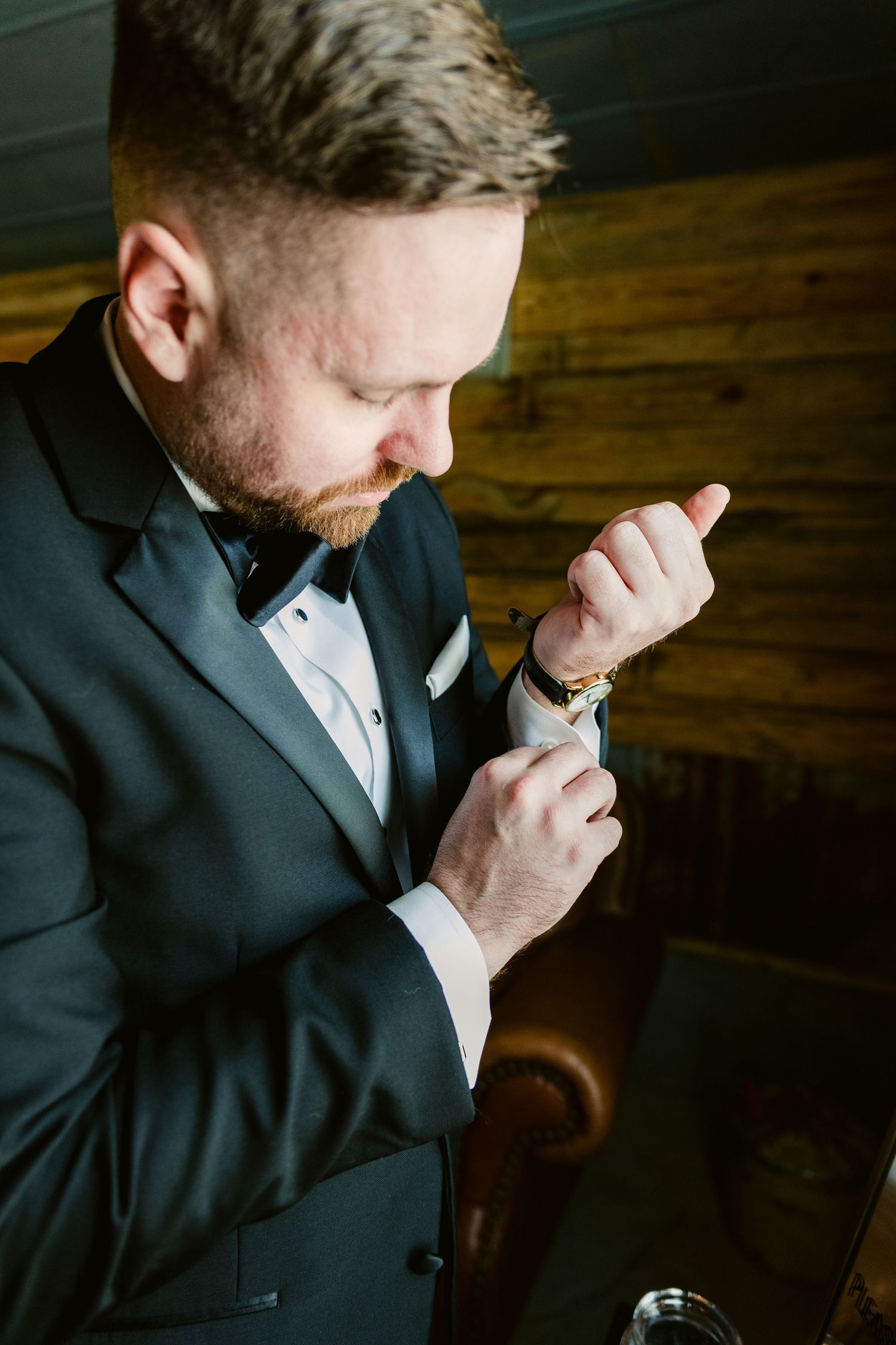 A person in a dark suit holds a cigarette while standing in moody lighting against a wooden wall.