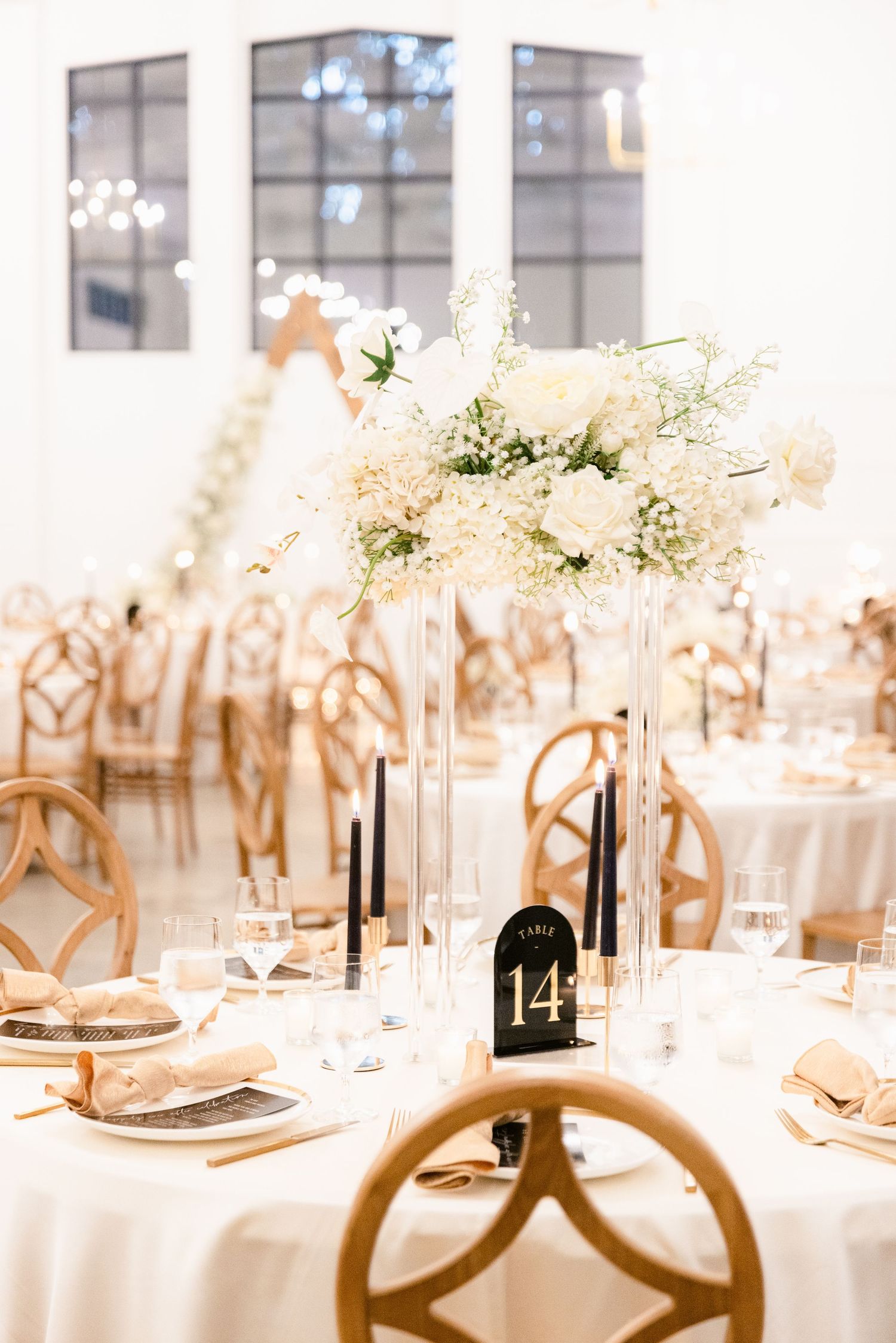 Elegant wedding reception table setting with white linens, floral centerpieces, and gold accents in a bright venue.