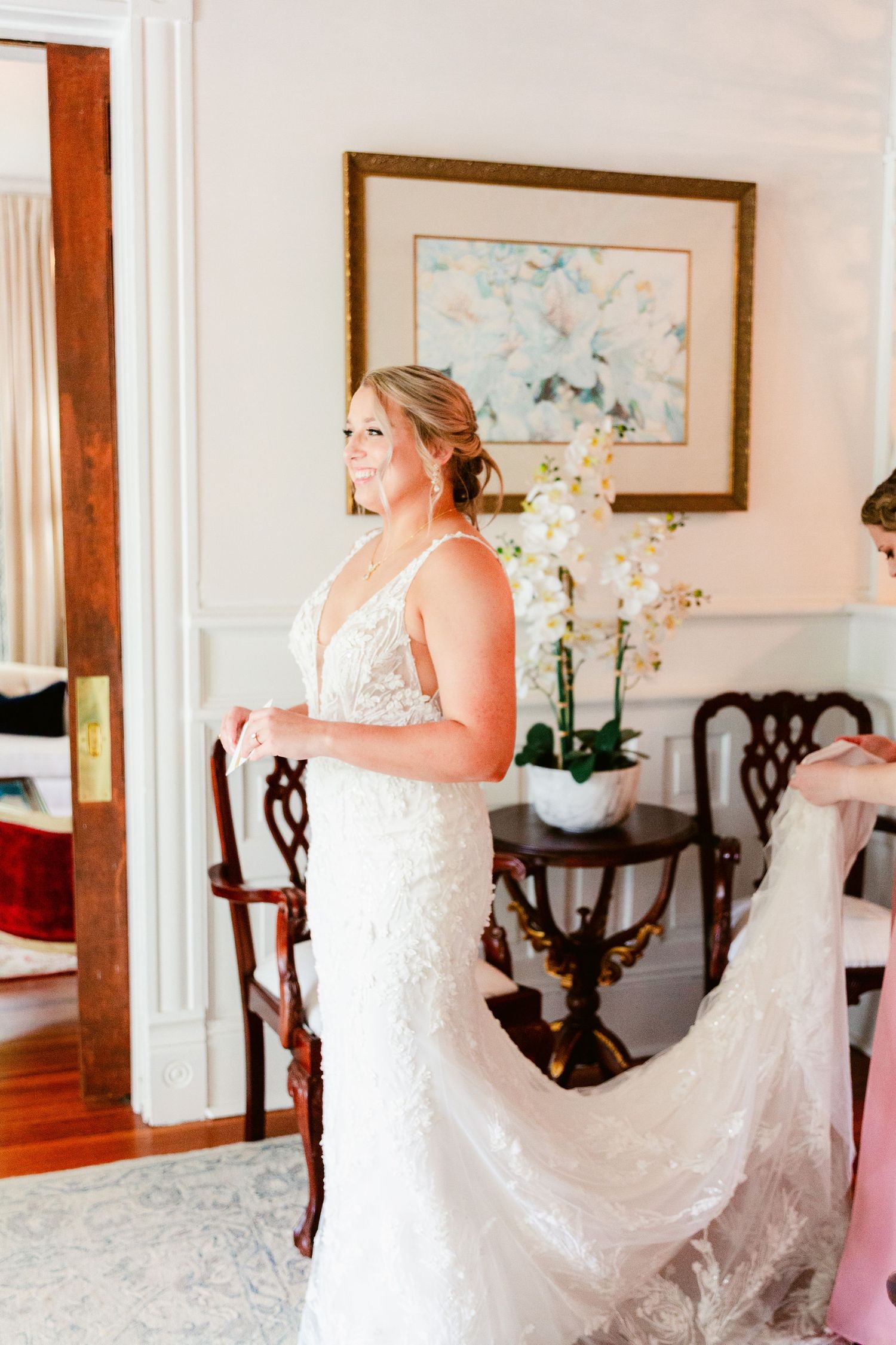 A bride wearing an elegant white lace wedding gown stands in an elegant room with hardwood floors and orchid decor.