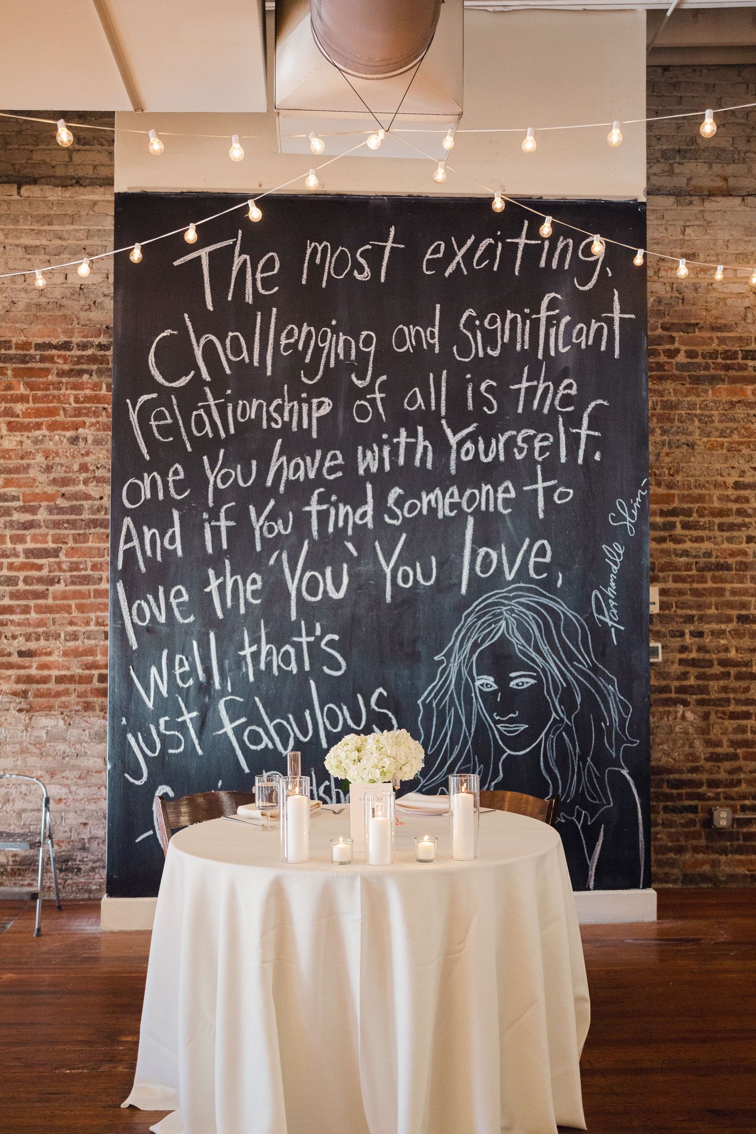 Wedding reception venue with chalkboard wall displaying inspirational quote and string lights in rustic brick setting.