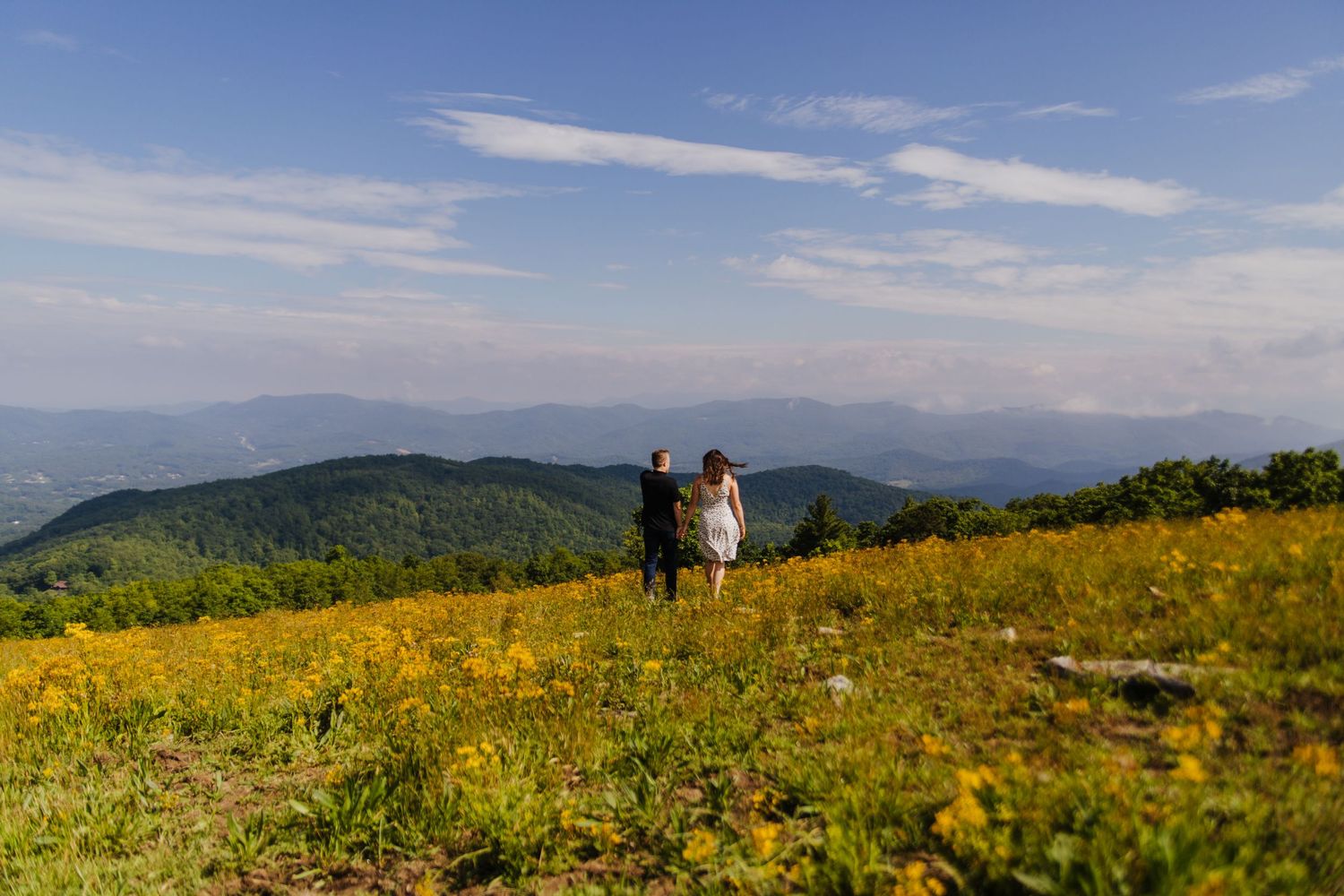 Couple stands together on a mountain summit overlooking rolling hills covered in yellow wildflowers.
