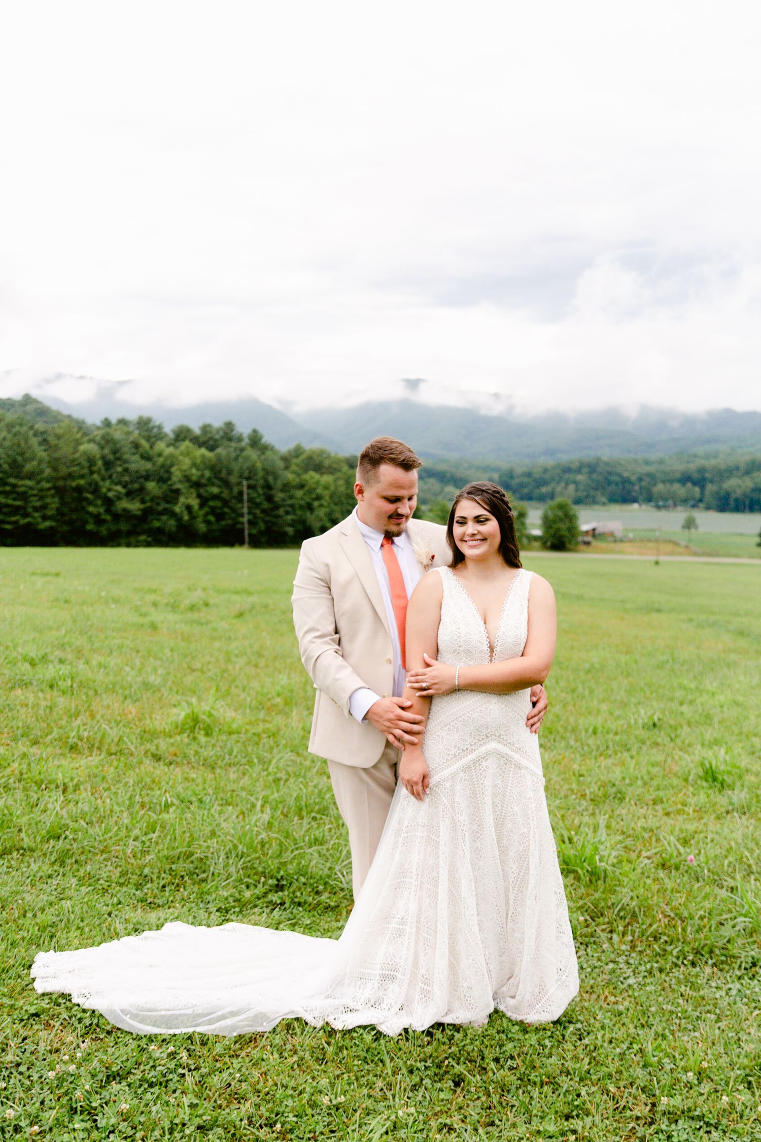 A couple in wedding attire sits together in a scenic mountain meadow with green grass and rolling hills in the background.