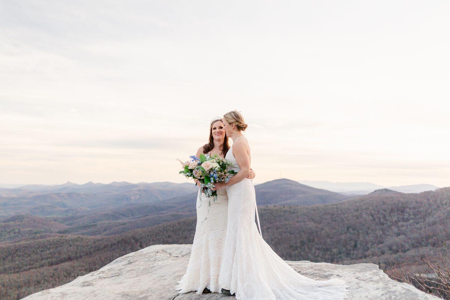 A couple in wedding attire stands on a mountain overlook with sweeping views of autumn foliage in the distance.