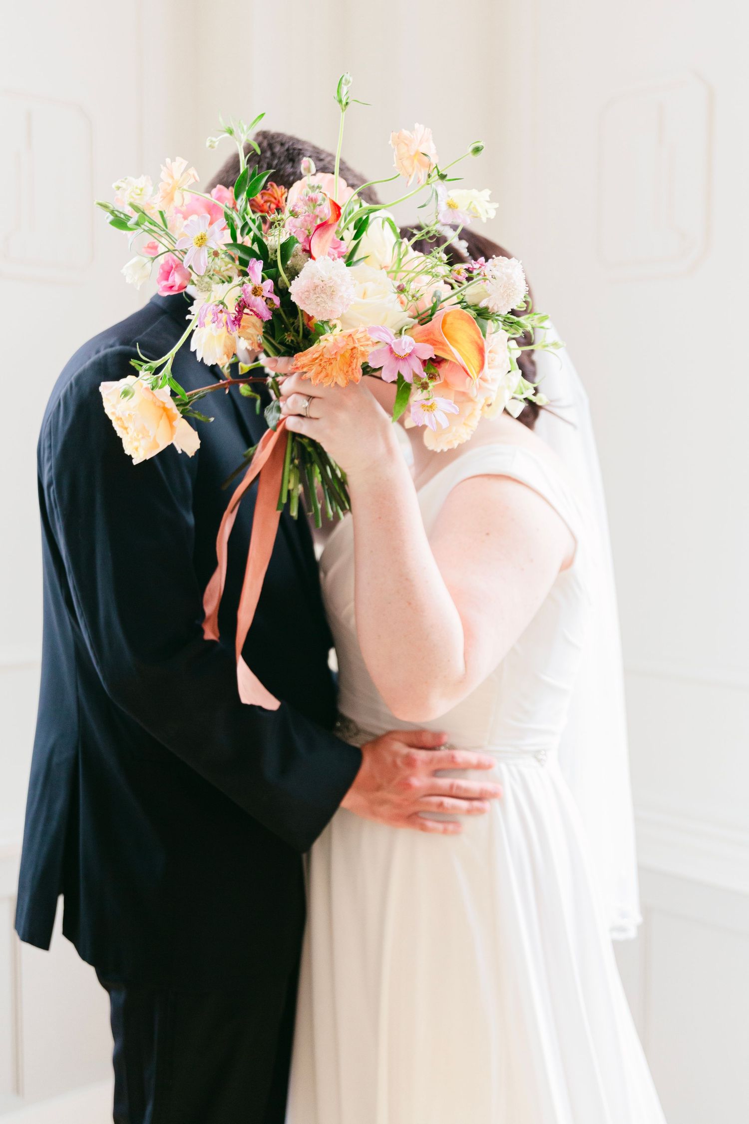 A romantic wedding couple embraces while holding a delicate peach and pink floral bouquet against a white backdrop.