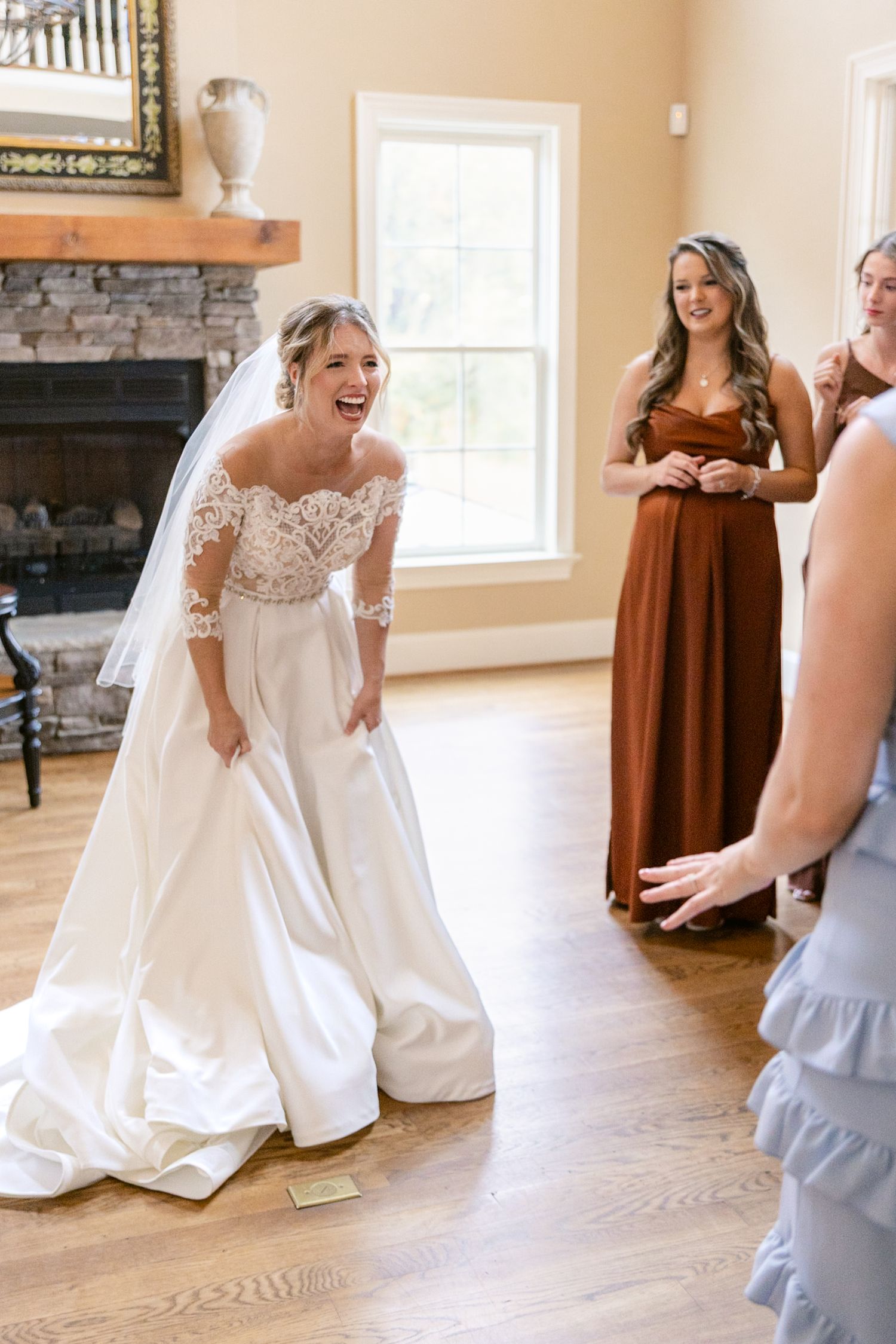 A bride in a white wedding gown reads a letter while bridesmaids in rust-colored dresses look on in a sunlit room.