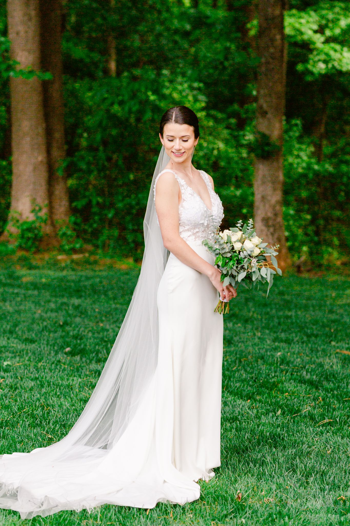 A bride in a flowing white gown poses gracefully on a lush green lawn surrounded by trees in a woodland setting.