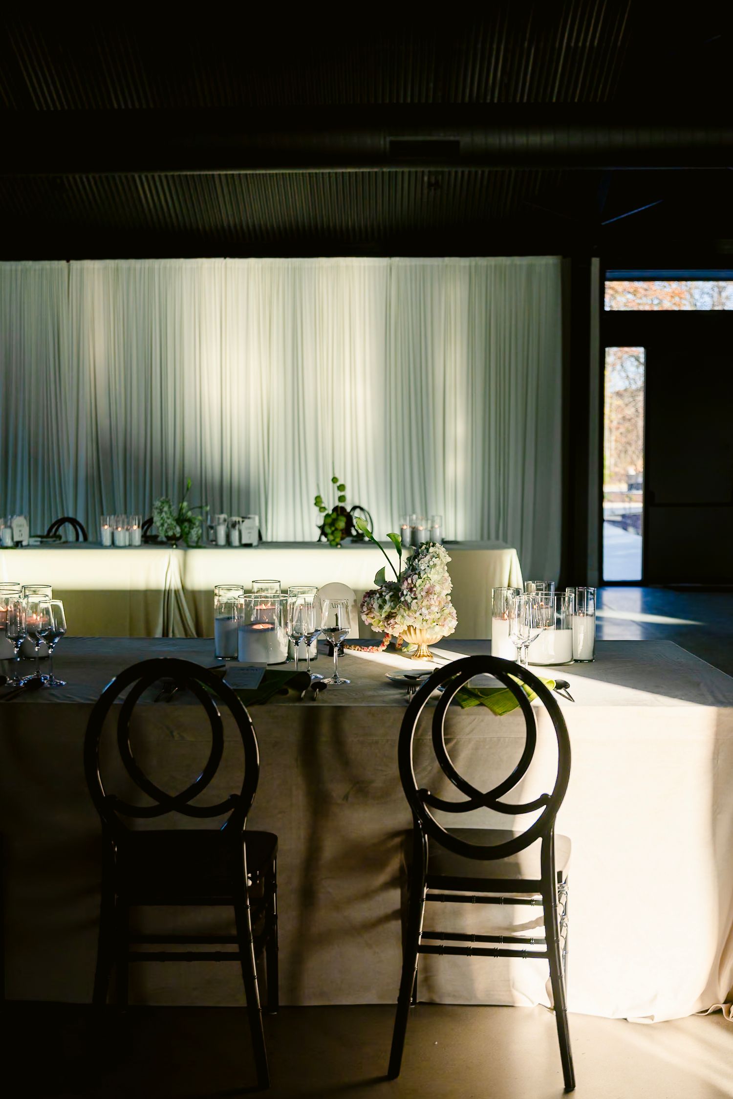 Elegant dining room tables with black chairs cast dramatic shadows in natural sunlight streaming through white curtains.