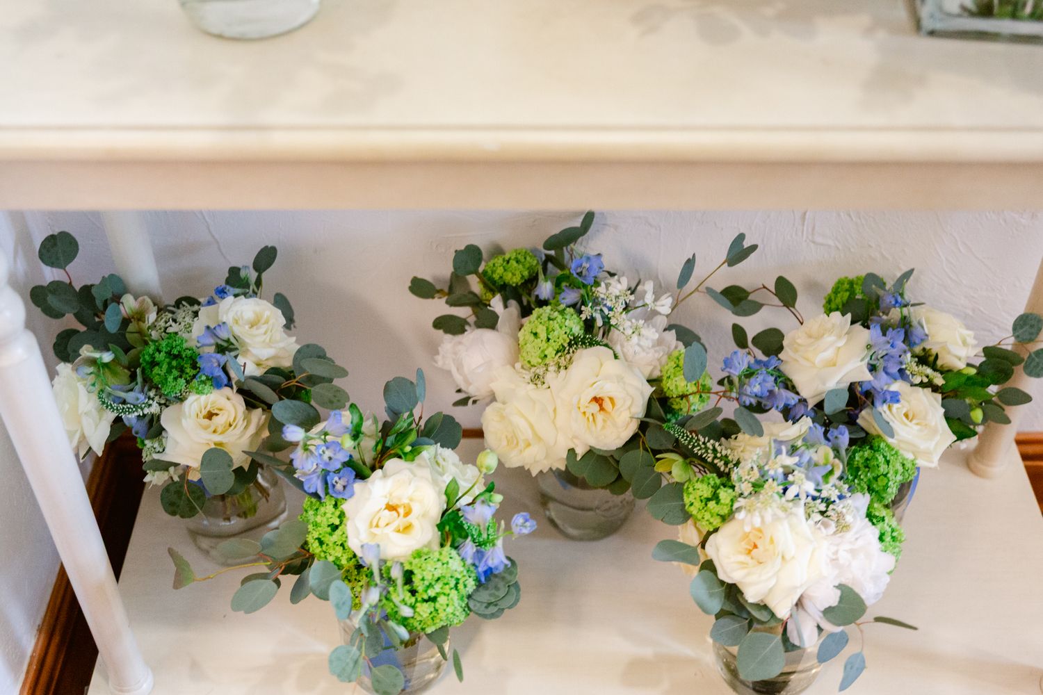 A row of elegant floral centerpieces with white roses and greenery in glass vases line a wedding reception table.