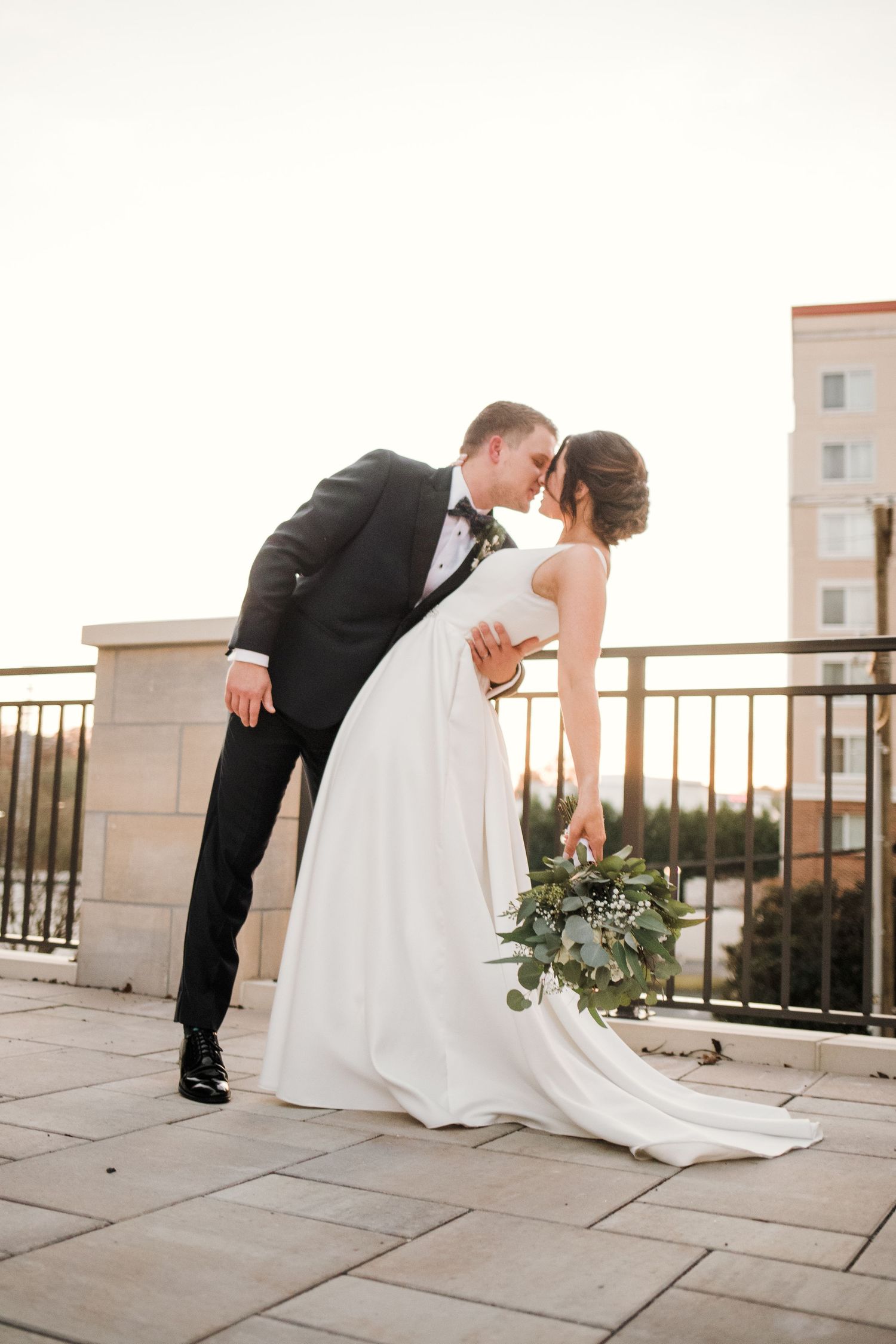 Classic wedding dip pose with bride holding elegant white and green bouquet against urban backdrop.