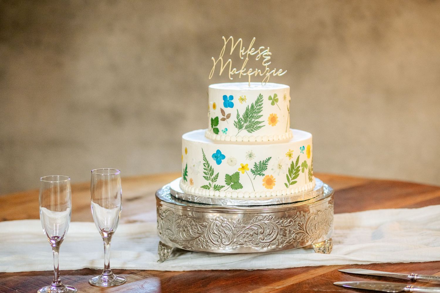 Two-tiered white wedding cake decorated with botanical designs on silver stand with champagne flutes.